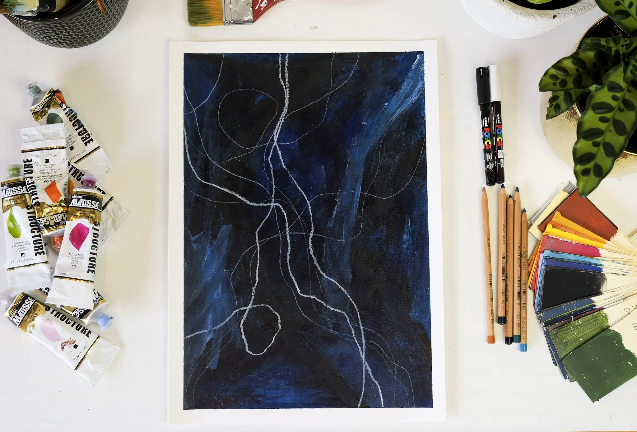 An abstract painting with a dark background and white and blue lines, surrounded by paint tubes, colored pencils, and color samples on a white table.