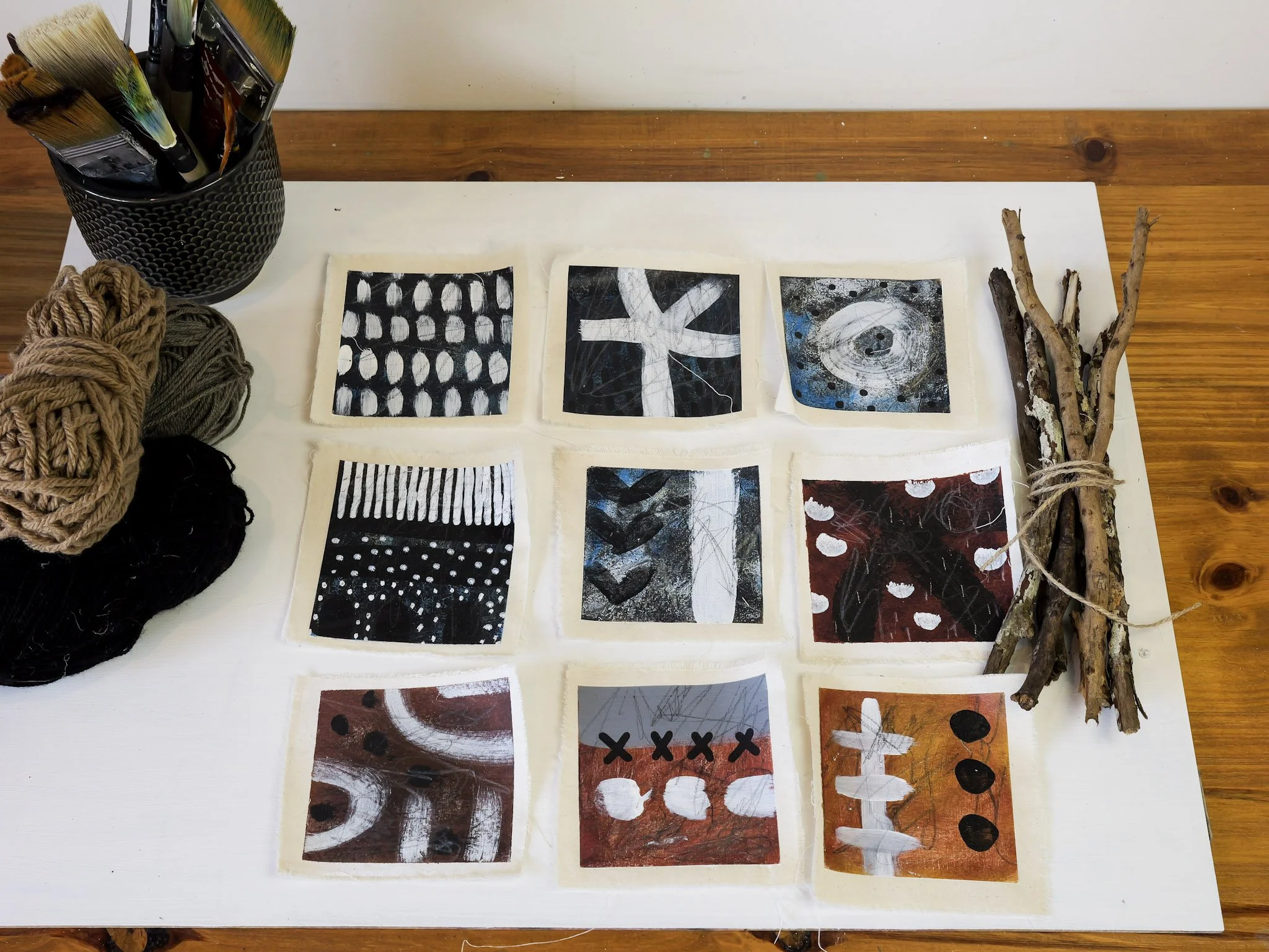Collection of nine small abstract paintings on fabric, arranged in a 3x3 grid, on a wooden table with art supplies and bundled sticks nearby.