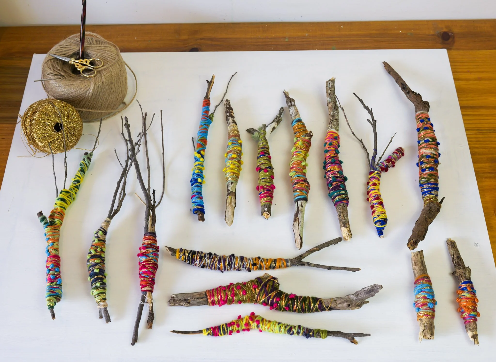 Several tree branches wrapped with colorful yarns arranged on a white surface, with balls of yarn and scissors at the top left corner.