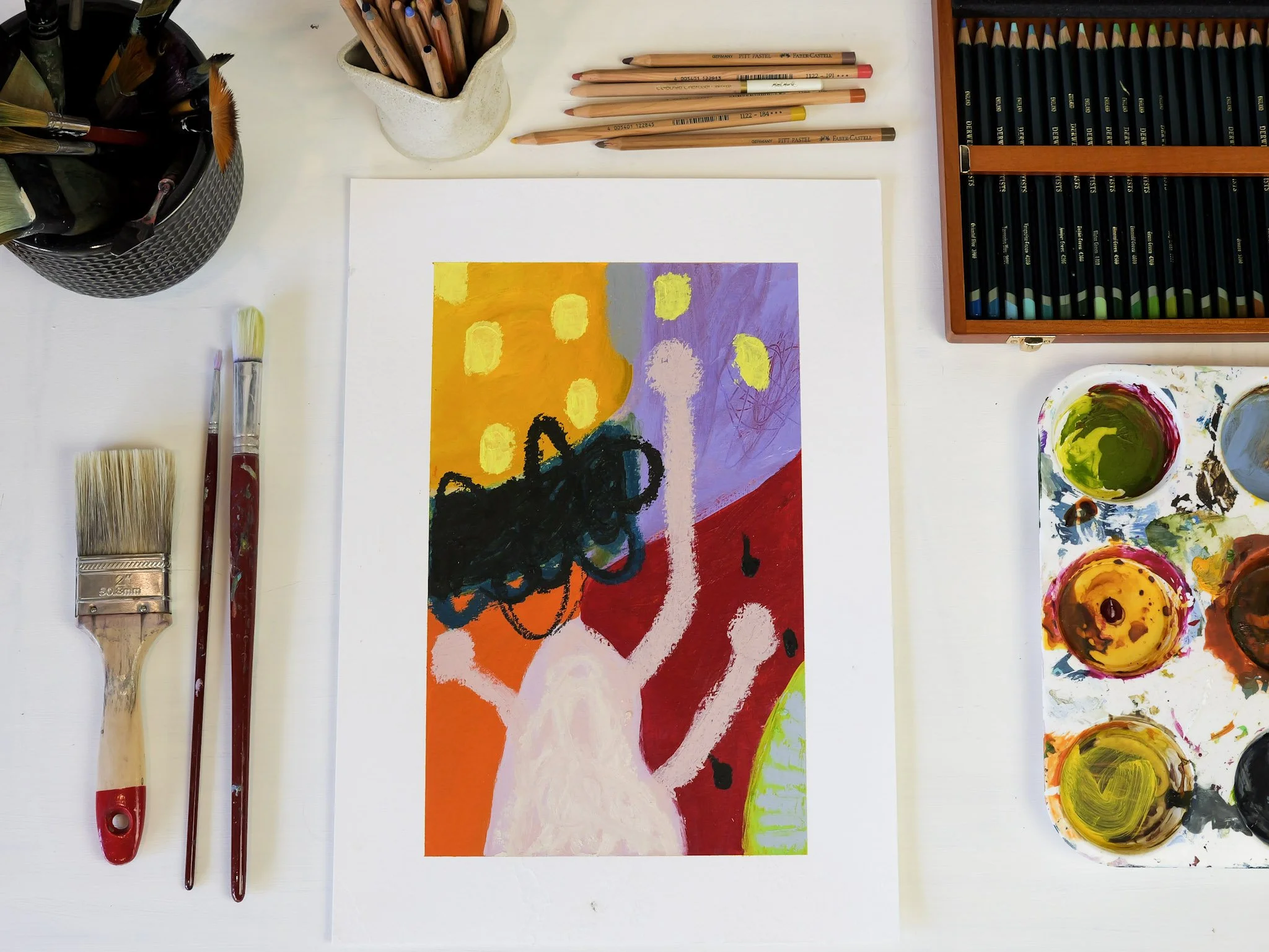 A colorful abstract painting in progress on paper, surrounded by paintbrushes, oil paints, and art supplies on a white table.