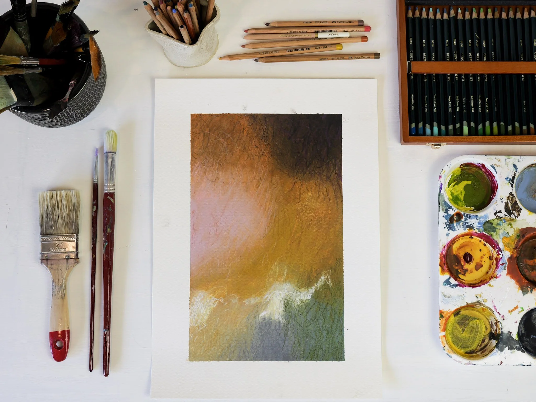 Watercolor painting in progress on paper, featuring warm earthy tones and abstract landscape with tree outlines, surrounded by brushes, colored pencils, and watercolor paints on a white table.