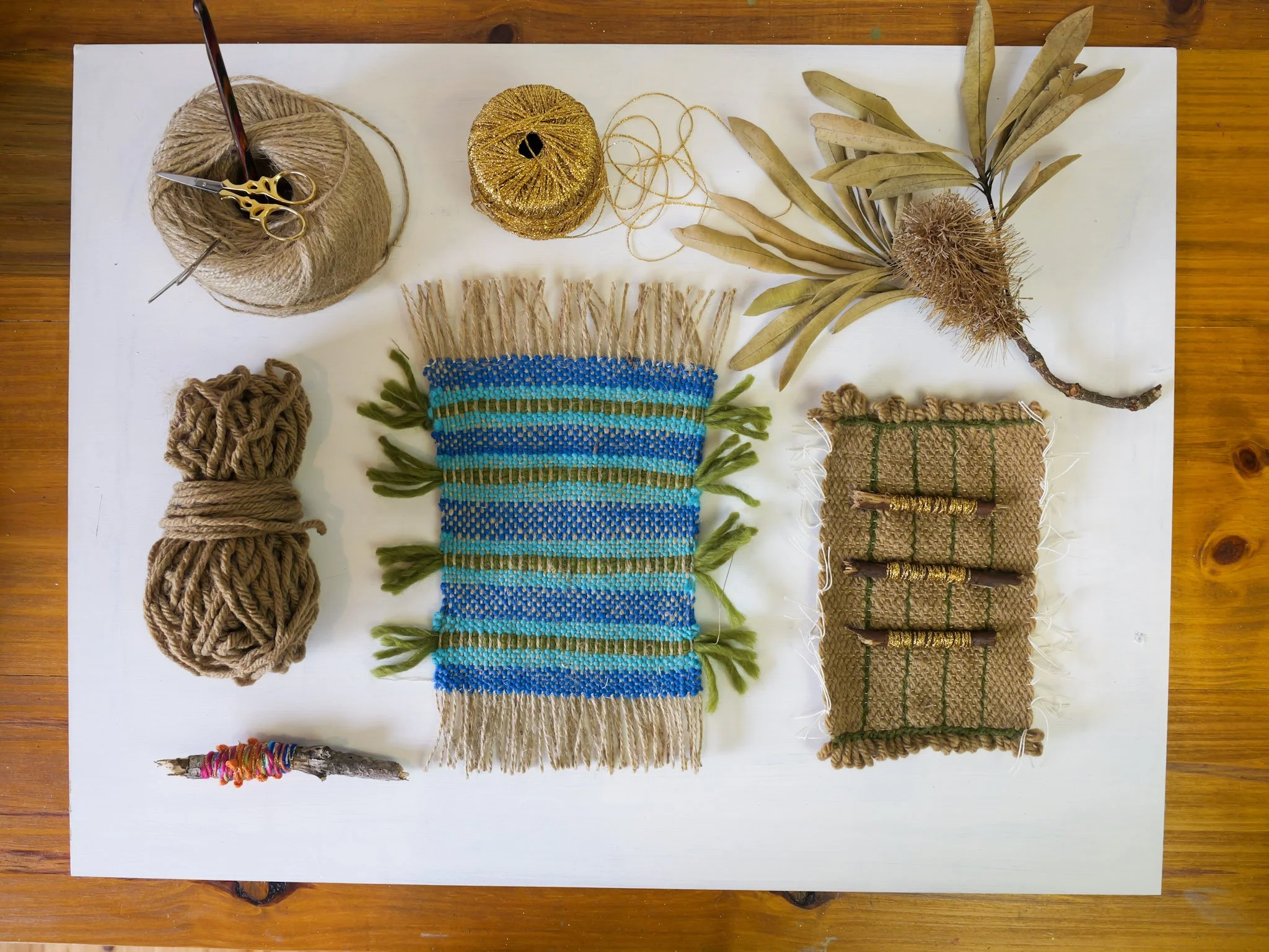 Flat lay of crafting supplies including two balls of yarn, a small branch wrapped with colorful thread, a woven textile with green and blue stripes, a bundle of dried leaves and a seed pod, a ball of beige yarn with scissors and a knitting needle, a 