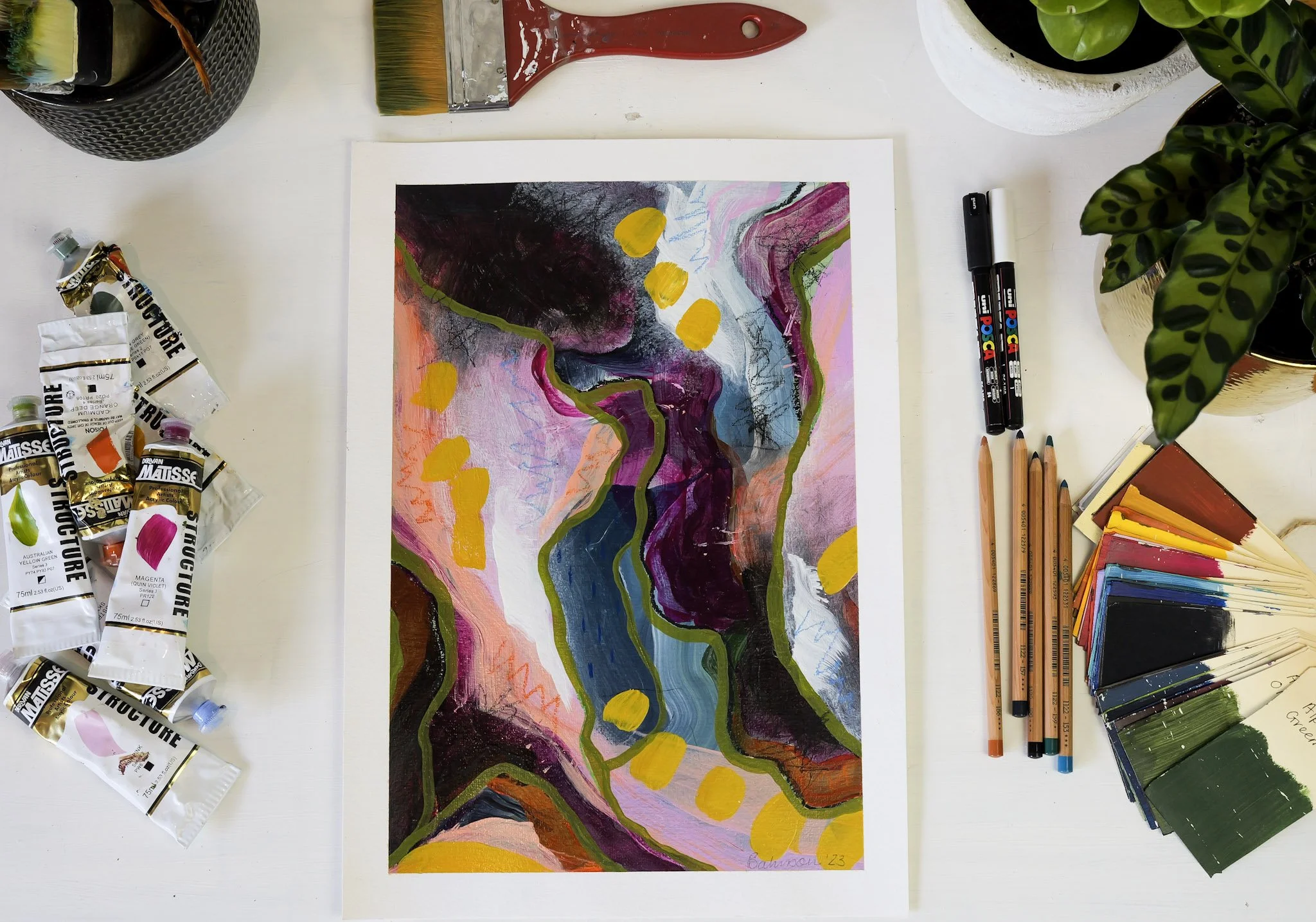 An abstract colorful painting on white paper surrounded by art supplies including paint tubes, paintbrushes, markers, and color swatches on a white table.
