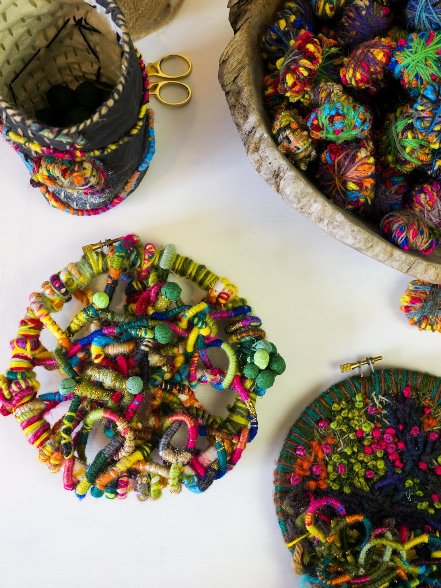 Colorful handmade crochet and knit jewelry on a white surface, with a basket of multicolored yarn, a pair of yellow scissors, and a tin container with black yarn balls.