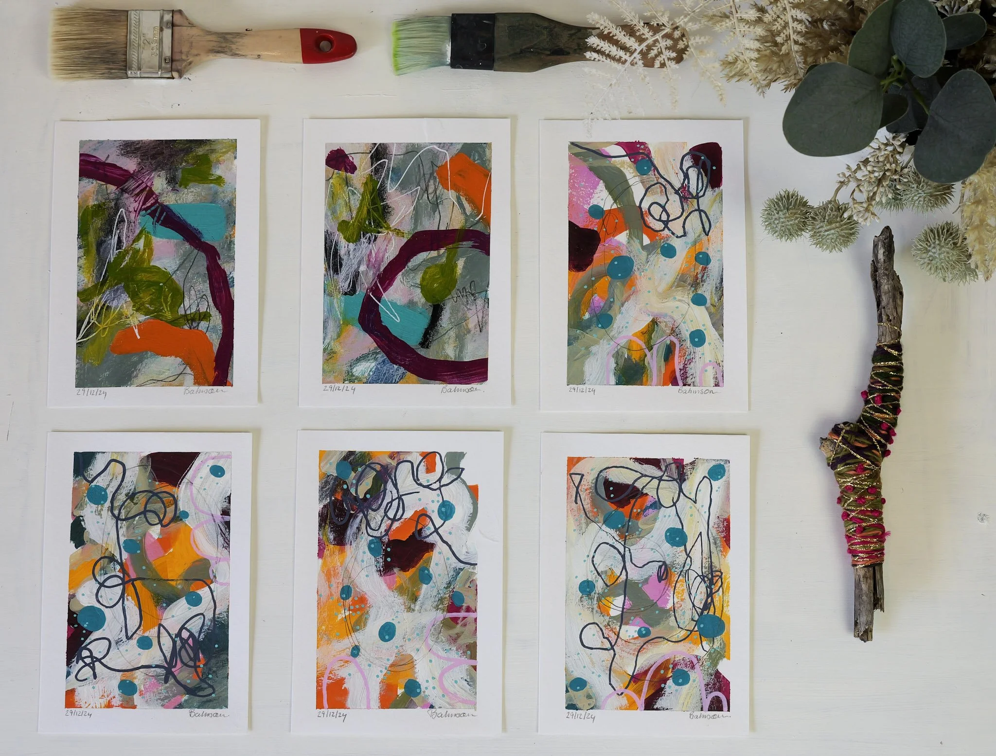 Six abstract paintings with vibrant colors and swirling lines on white paper, each signed "Balmoon" and dated "29/12/24," arranged in two rows of three on a white table. To the right of the paintings, there is a decorative wand wrapped with colorful 