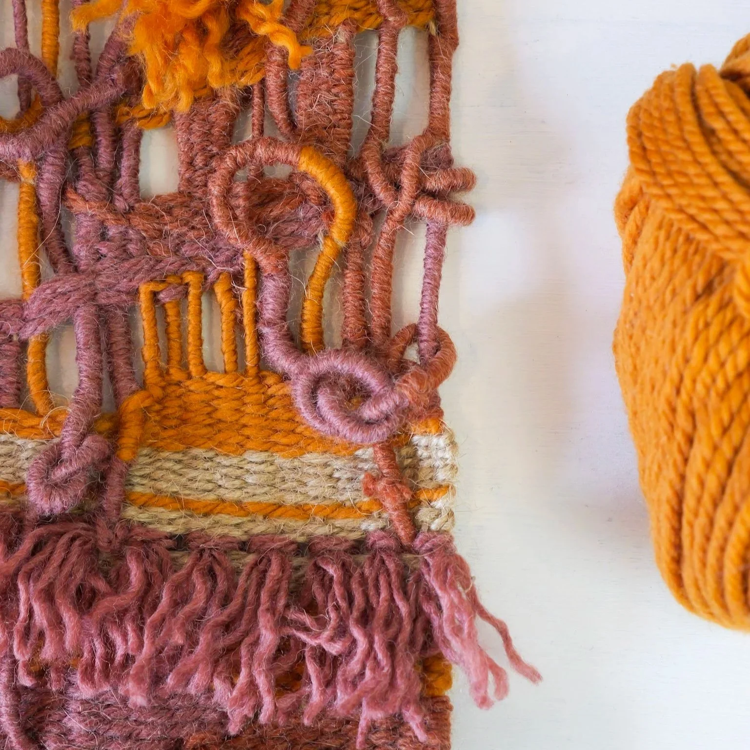 Close-up of a woven textile with pink and orange yarns with a fringed edge, and a large ball of orange yarn to the right.