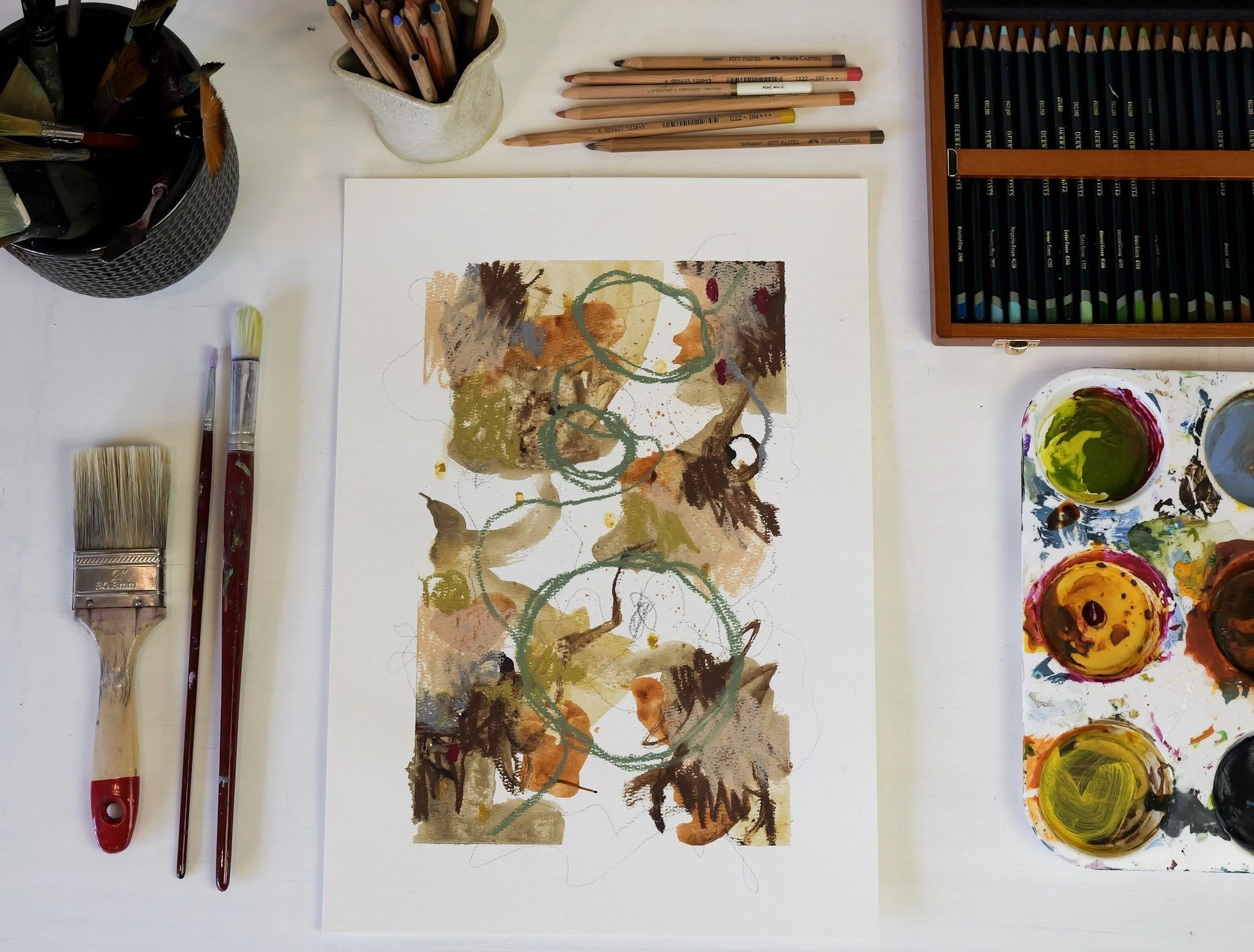 Abstract watercolor painting on white paper with circular shapes and earthy tones, surrounded by art supplies including brushes, colored pencils, and paint palettes.