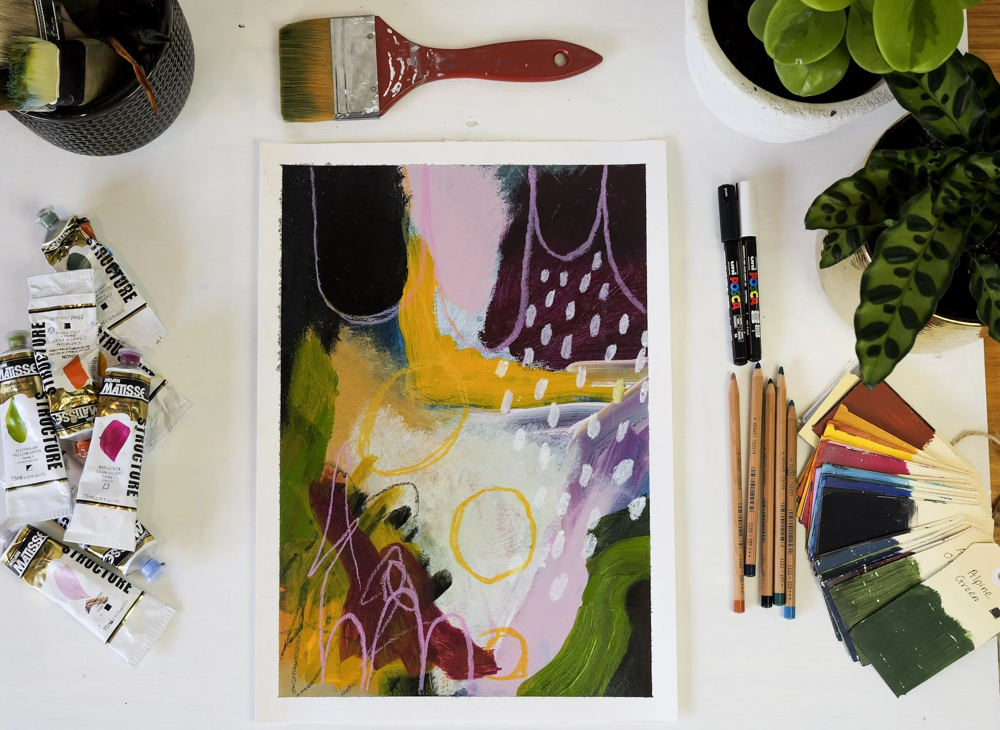 An abstract colorful painting on paper, surrounded by paint supplies, markers, colored pencils, paint tubes, paint swatches, and potted plants on a white table.