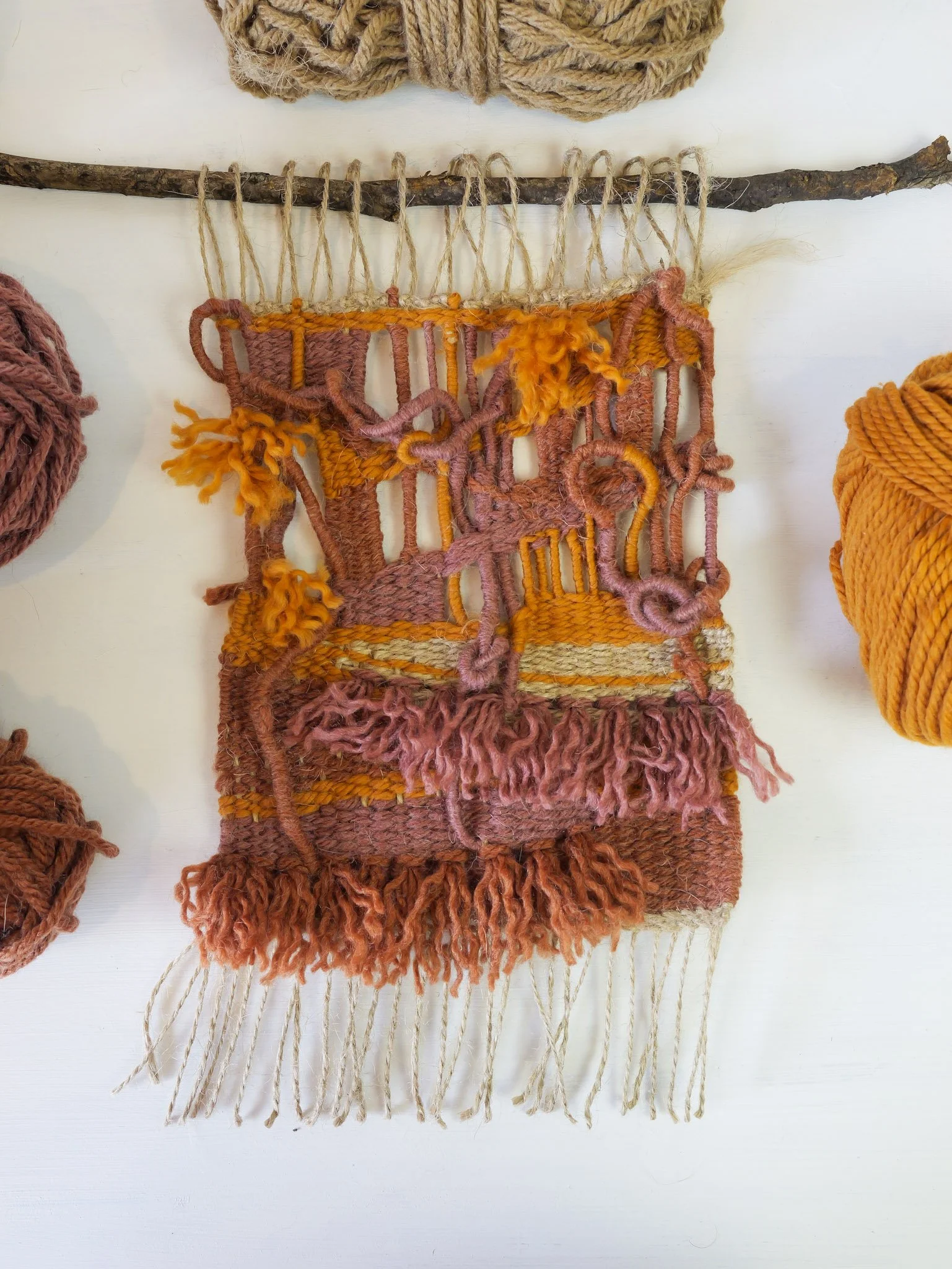 A woven textile artwork with abstract patterns in pink, orange, and yellow yarn, hanging from a branch.
