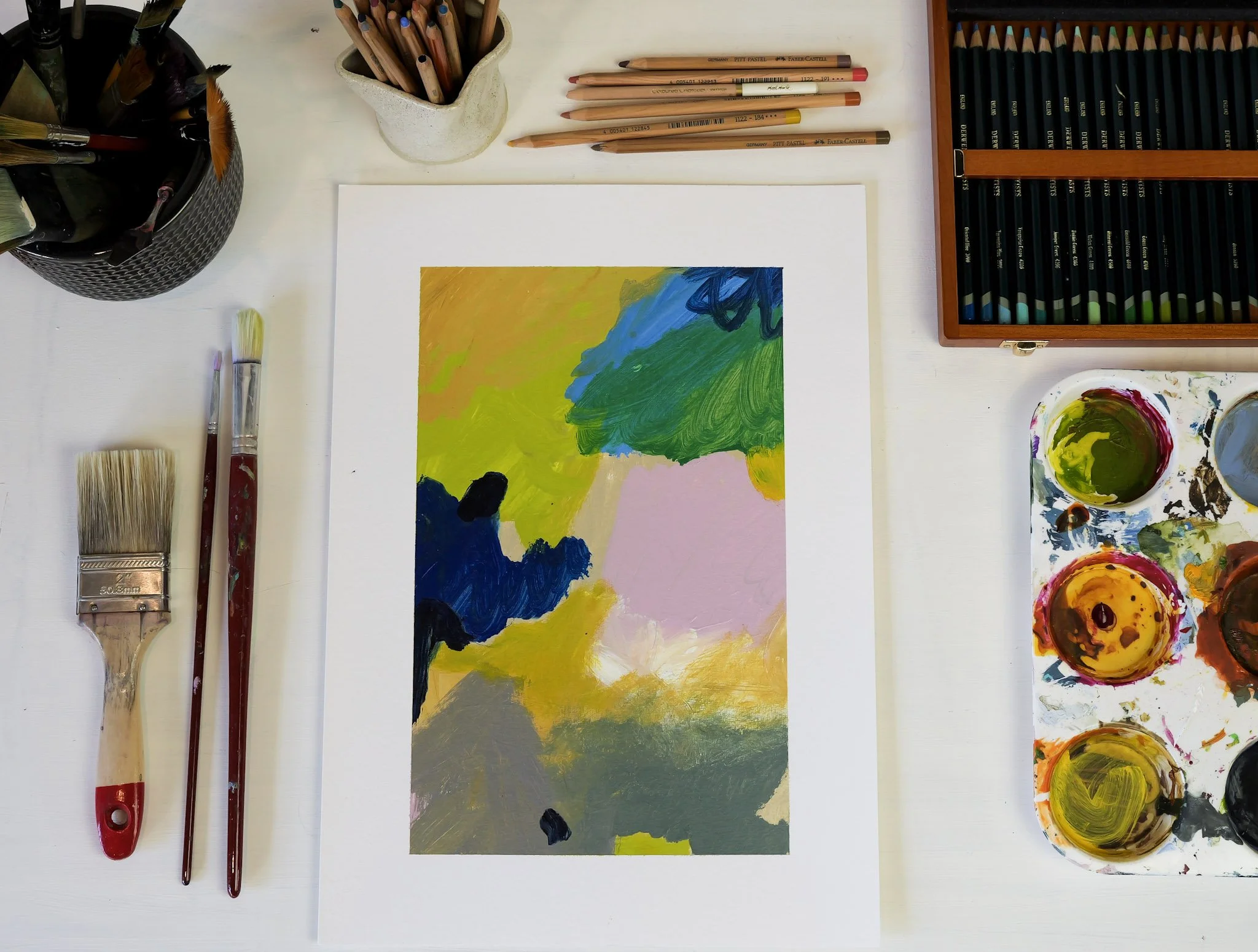 Abstract landscape painting with vibrant colors, including green, blue, yellow, pink, and gray, on white paper. Art supplies around it include paintbrushes, watercolor palette with yellow, green, and red paints, colored pencils, and jars of brushes a