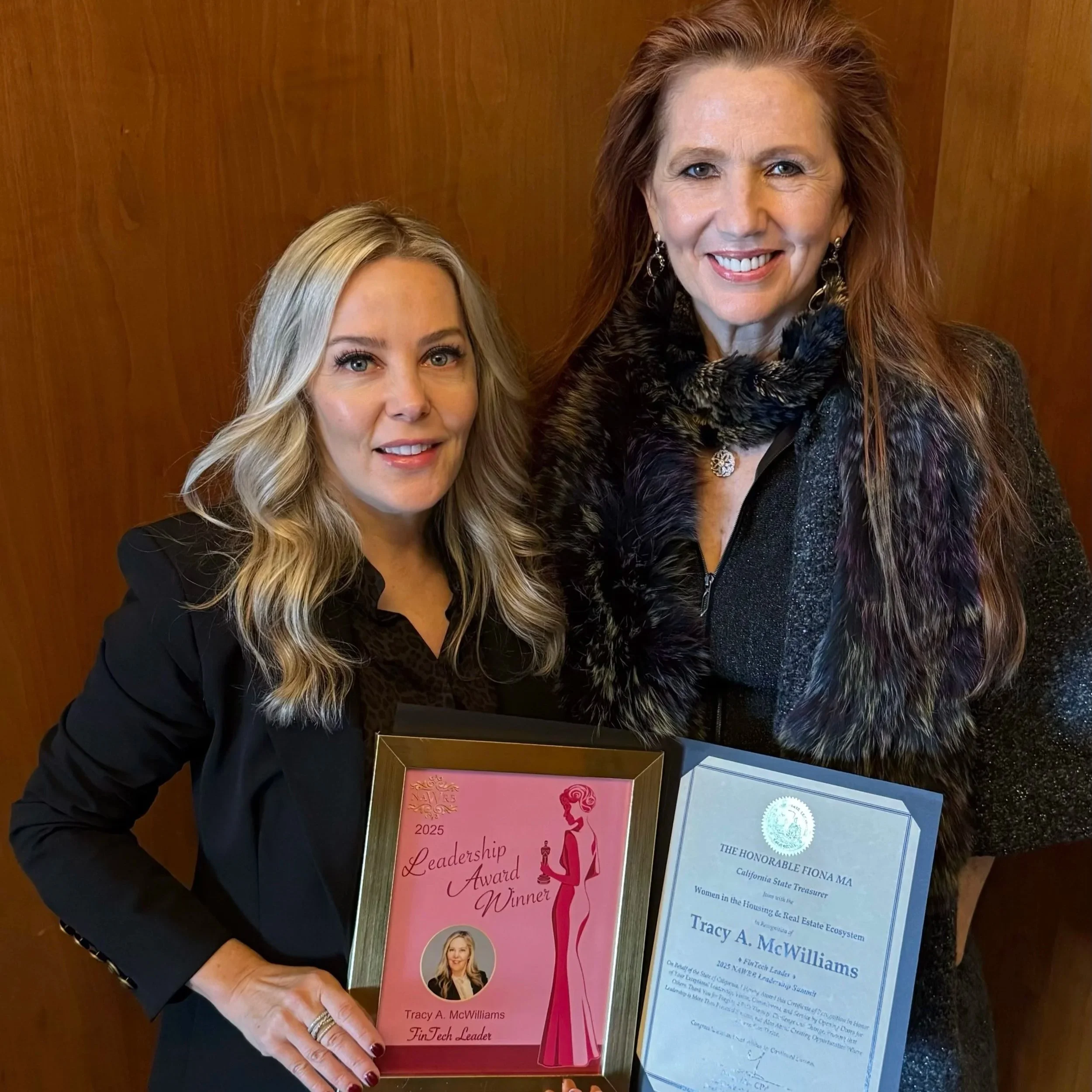 Two women standing together, holding awards and certificates, smiling at the camera. One woman has long, wavy blonde hair and wears a black blazer; the other has long red hair, wears earrings, a black dress with a furry black shawl, and a necklace. The awards indicate recognition in leadership and housing and real estate.