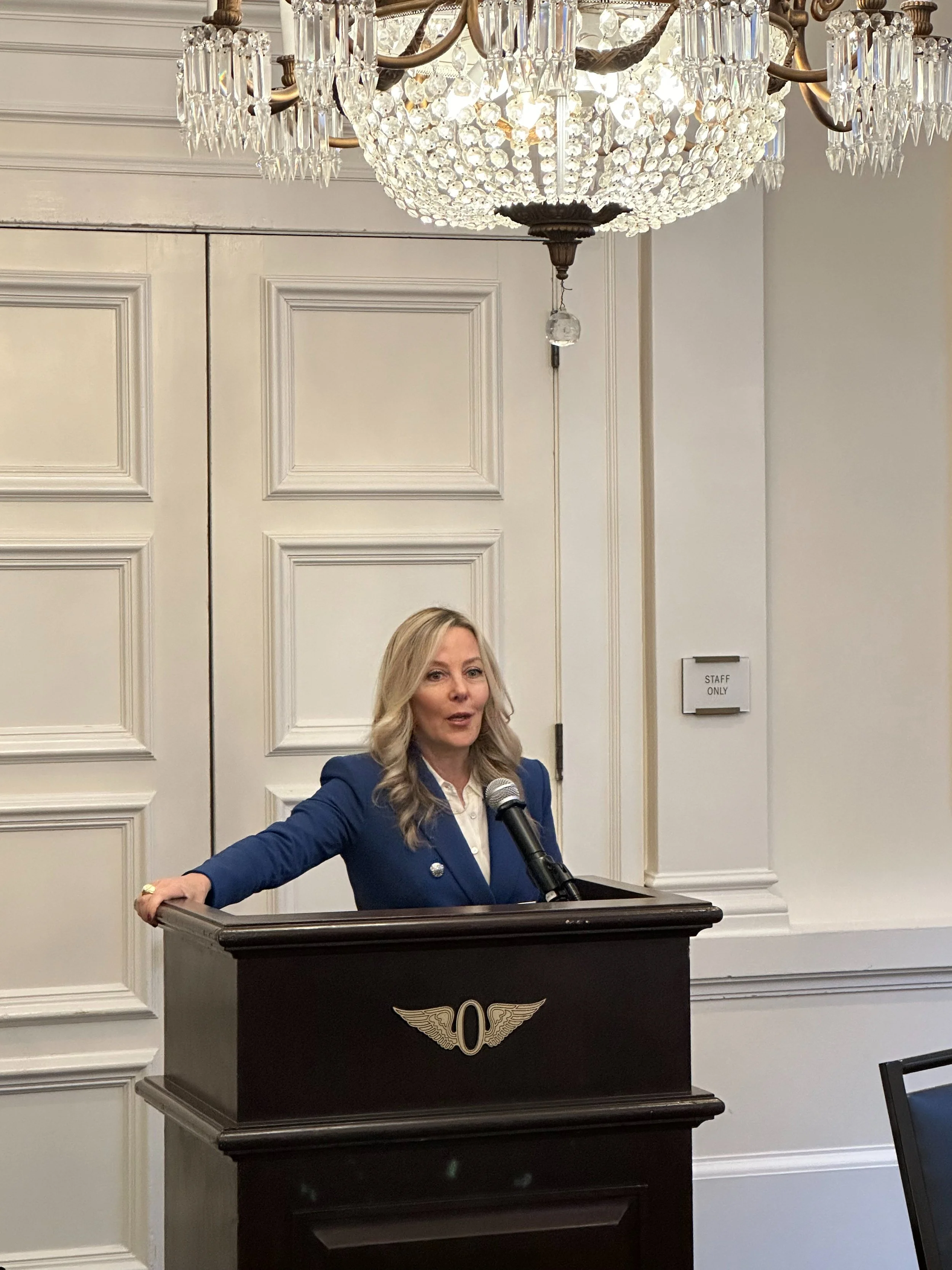 A woman in a blue blazer standing at a podium with a microphone, indoors in a room with white paneled walls and a large crystal chandelier overhead.