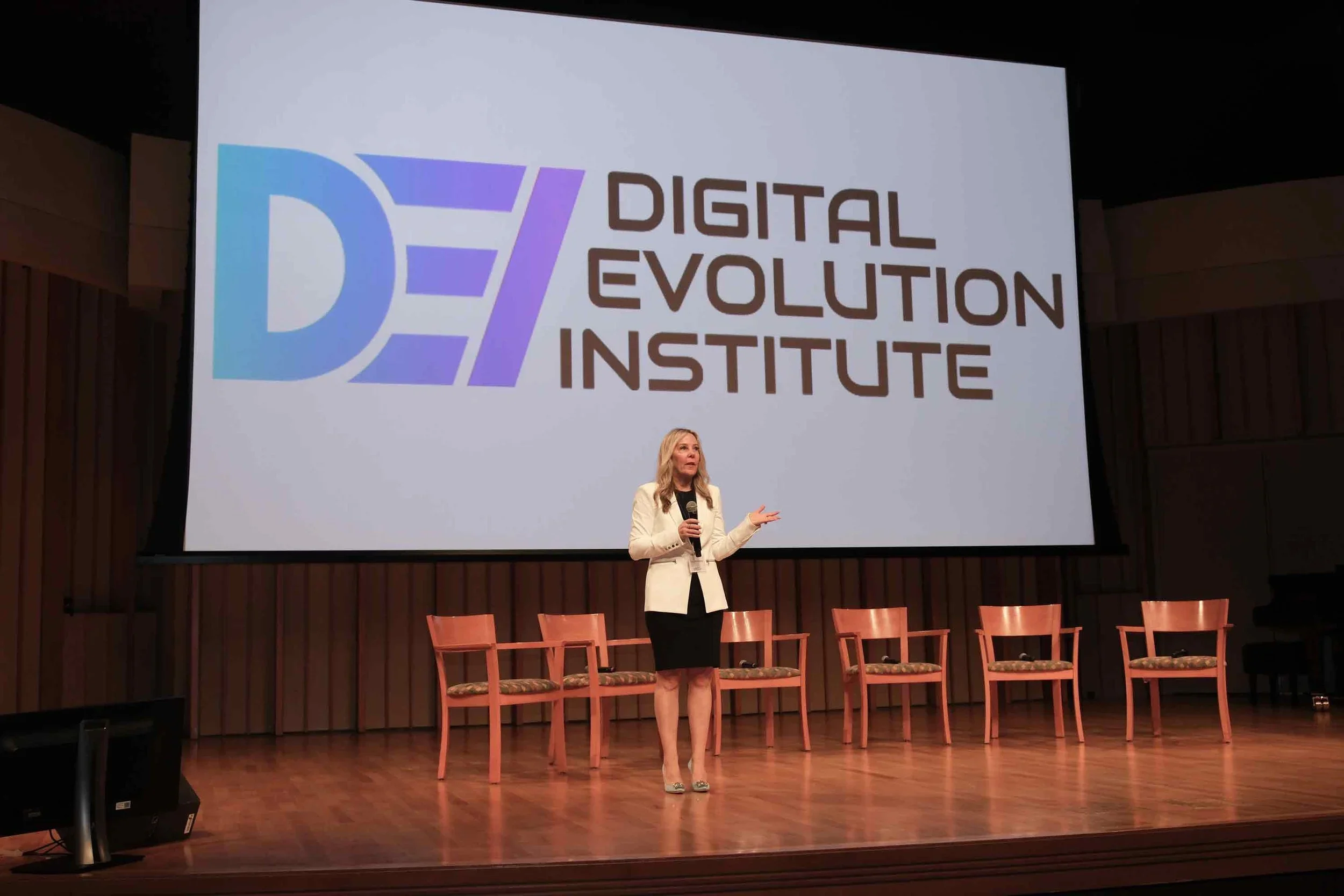 A woman standing on a stage holding a microphone, speaking in front of a large screen that displays the logo and name of the Digital Evolution Institute. The stage has six empty chairs arranged in a row behind her.