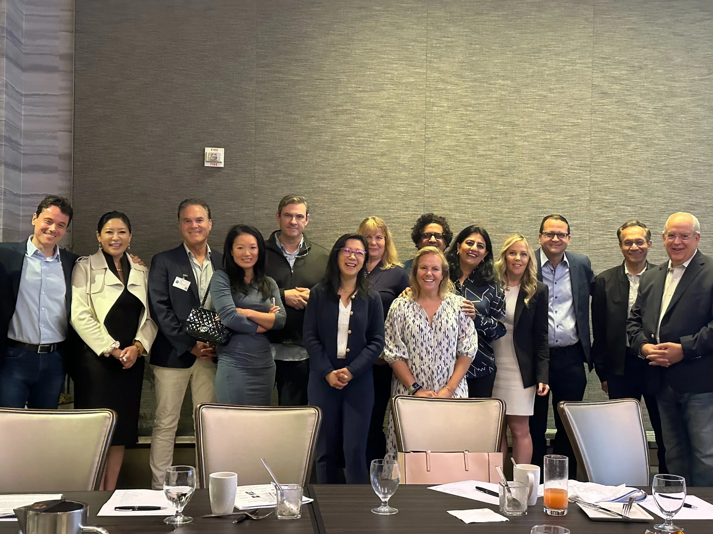 Group of 14 people standing in a line, smiling, in a conference room with a tan wall and a table in the foreground with glasses, cups, and papers.