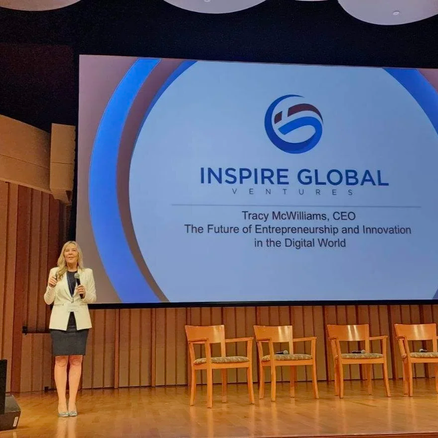 A woman in a beige blazer and dark skirt standing on a stage holding a microphone, next to a large screen displaying a presentation titled 'Inspire Global Ventures' with the logo, and the name Tracy McWilliams, CEO, with a subtitle about entrepreneurship and innovation in digital world. There are four empty chairs on the stage.