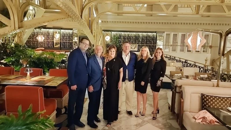 Group of six people dressed in formal attire standing inside a restaurant with modern decor and warm lighting.