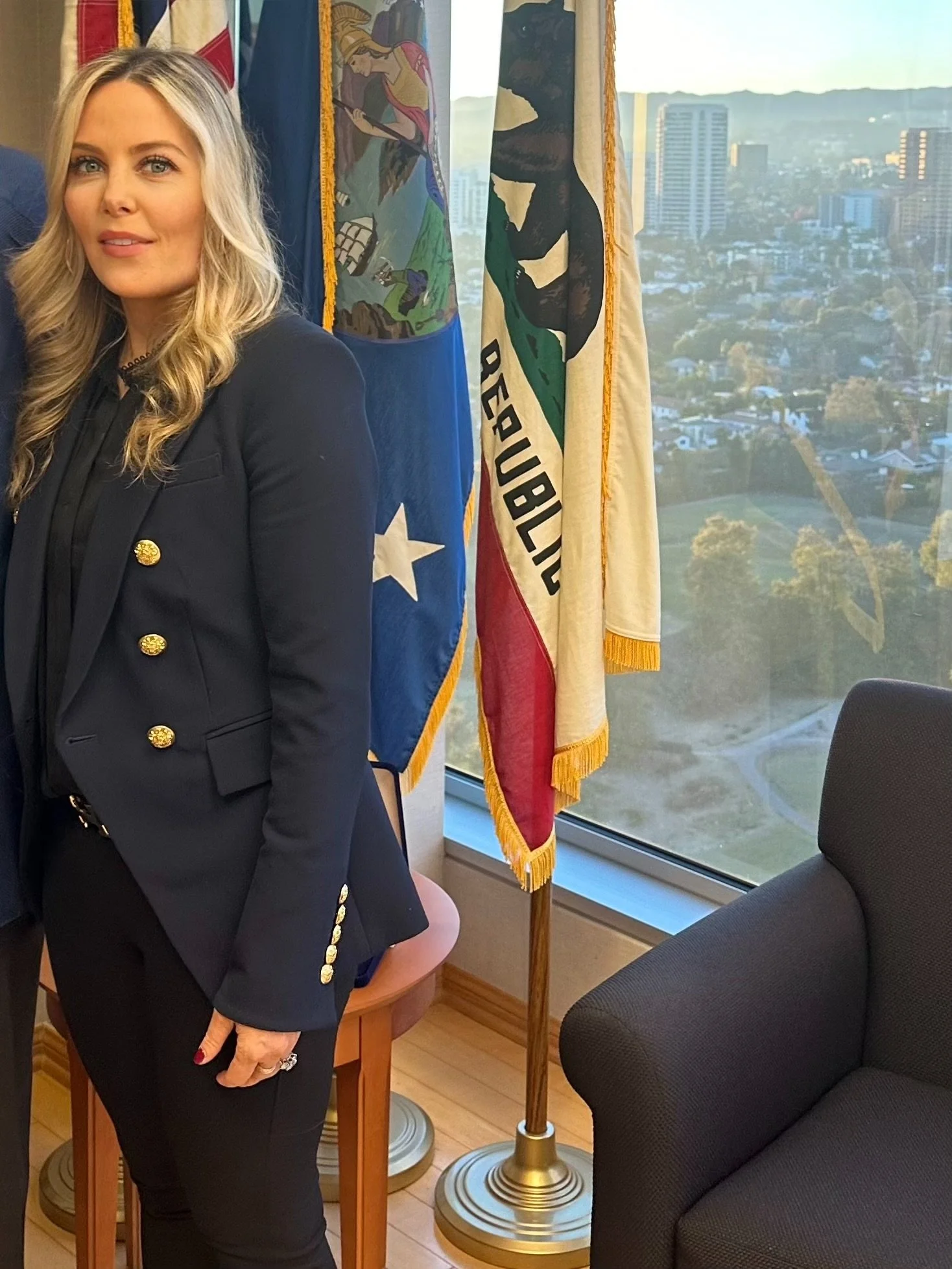 A woman with long blonde hair wearing a dark blazer with gold buttons standing next to a flag of California in a room with large windows overlooking a cityscape.