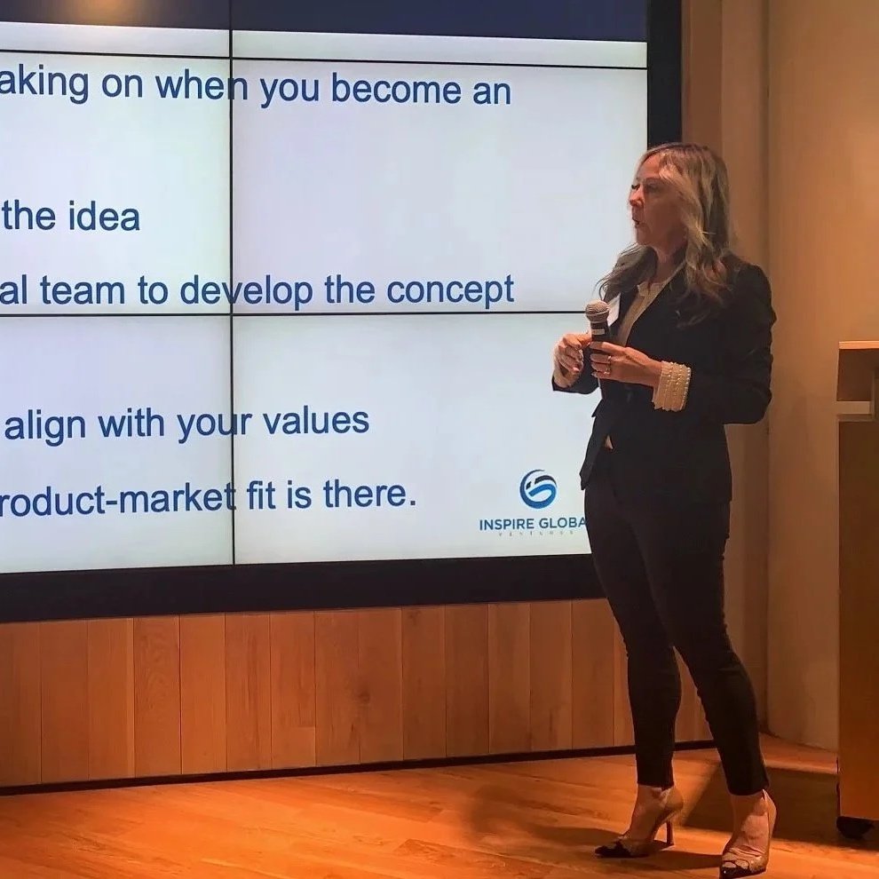 Woman in black suit with beige cuffs presenting at a conference, holding a microphone, standing in front of a large screen displaying a slide with text and a logo.