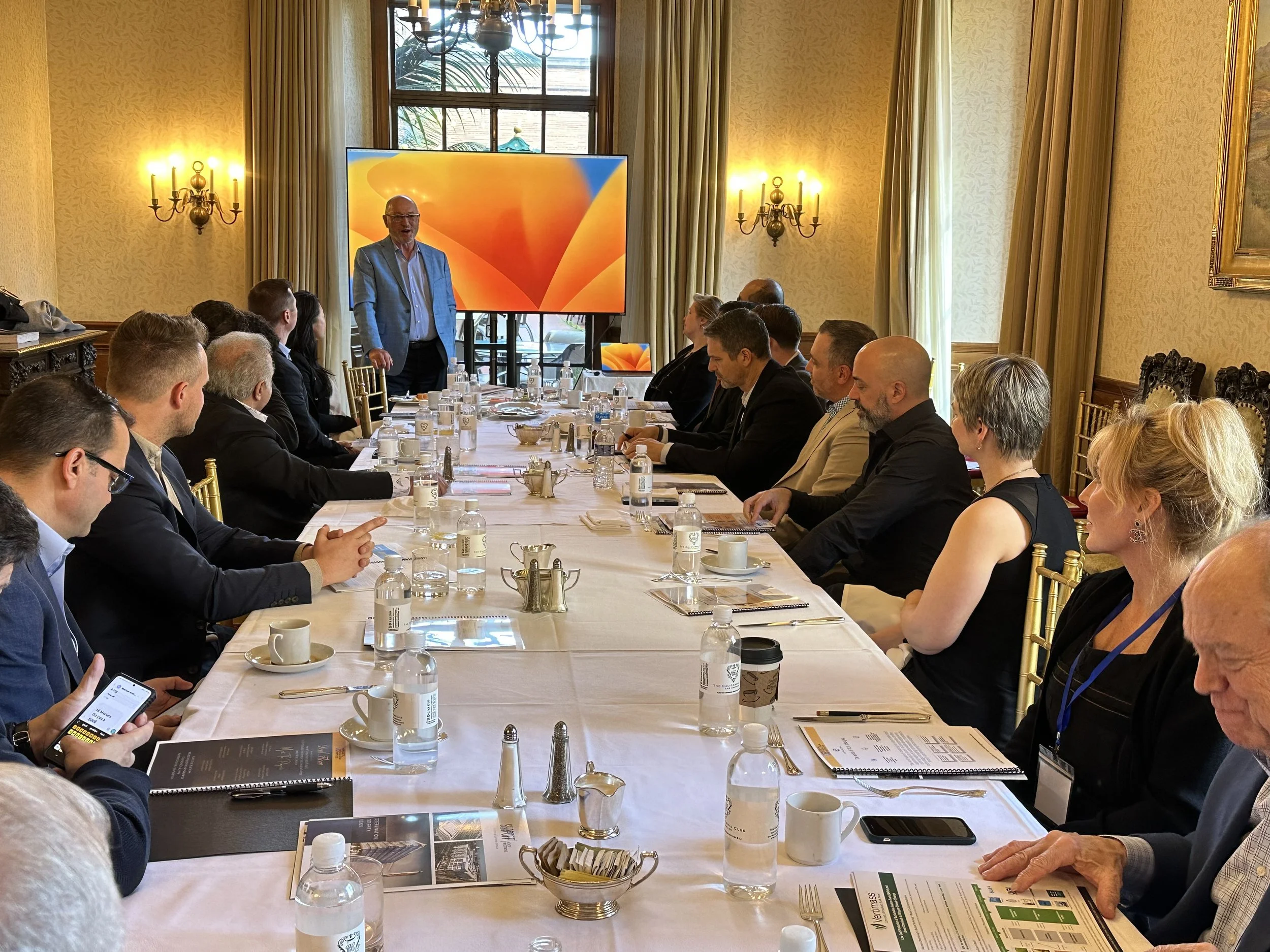 A business meeting in a formal conference room with attendees seated around a long table, listening to a speaker standing at the front, with a large screen behind him displaying vibrant orange graphics.