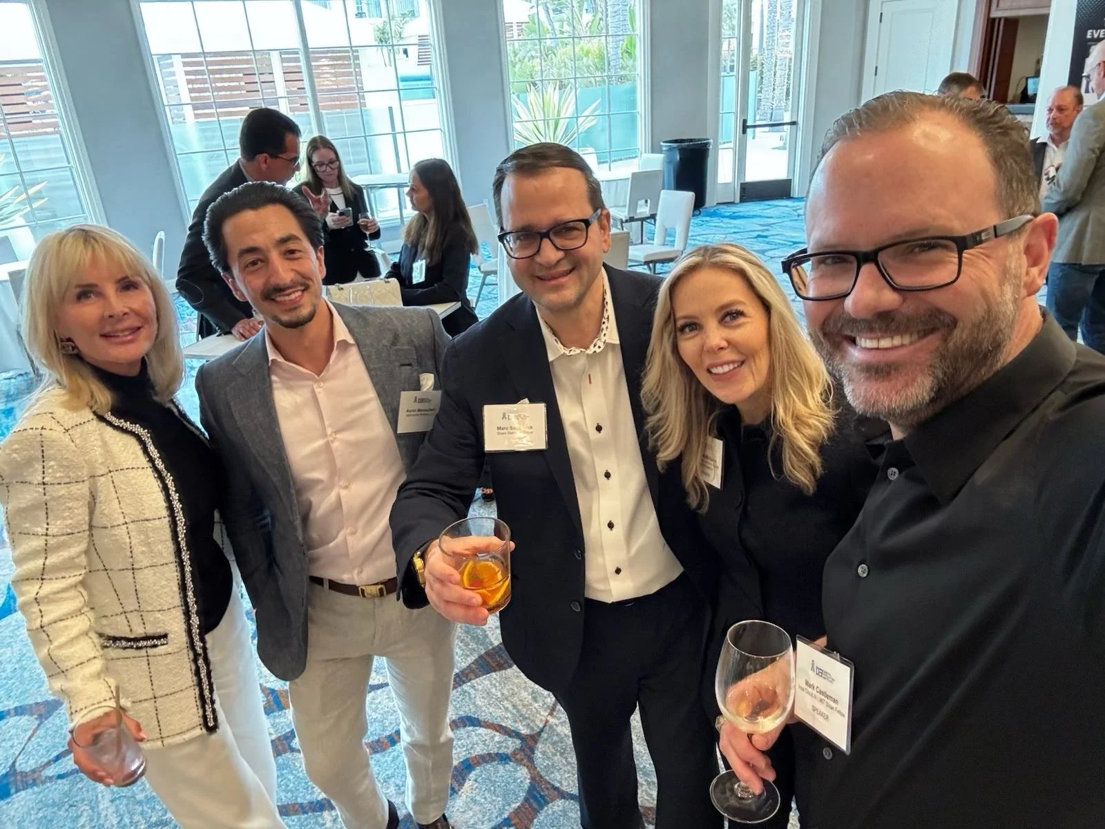 A group of five people at a social event, smiling and holding drinks, with others in the background near large windows and blue carpeted floor.