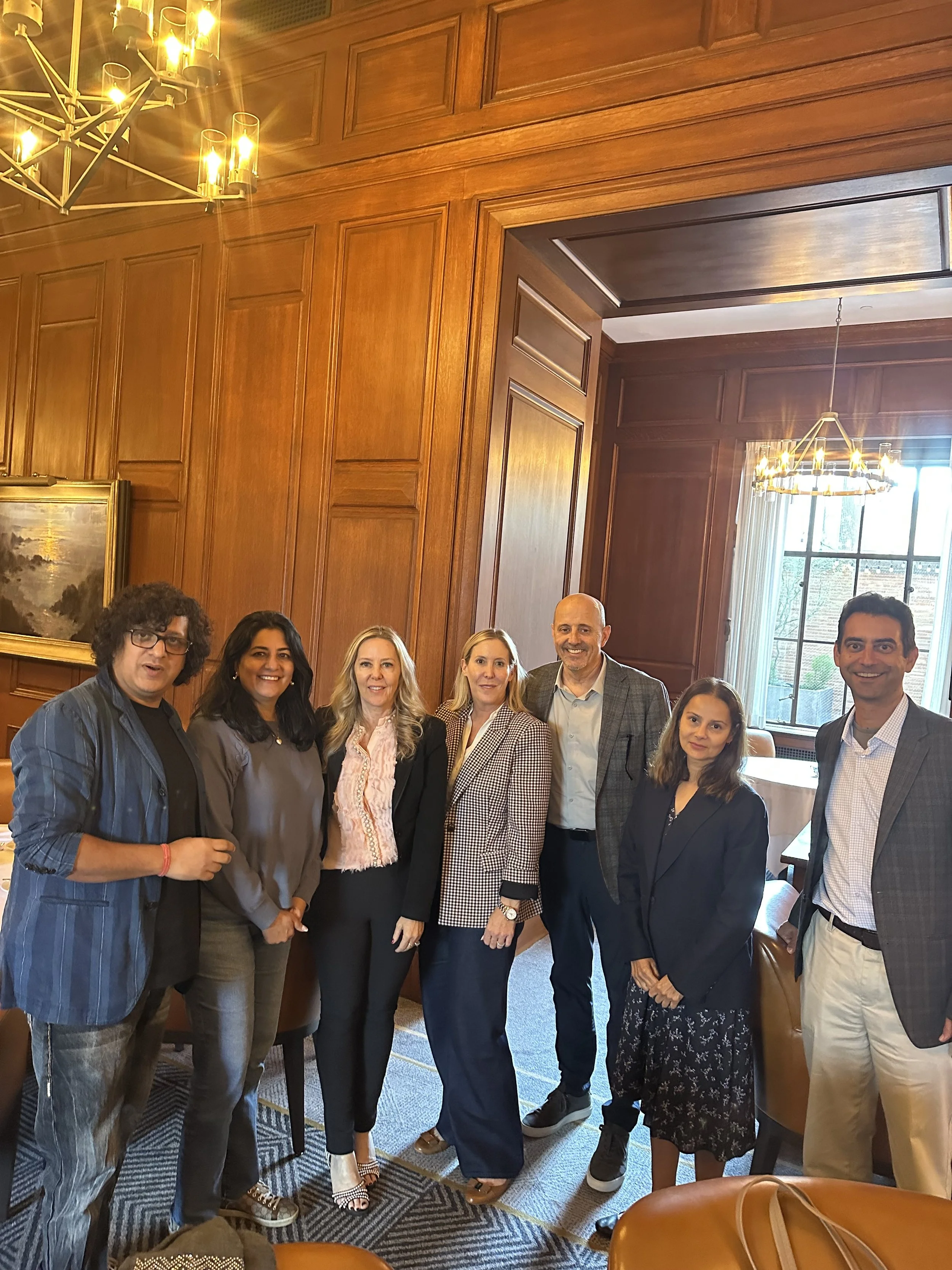 Group of seven diverse people, five women and two men, smiling and posing in a wood-paneled room with large windows and chandeliers.