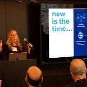 A woman presenting at a conference, standing next to a large screen displaying the text 'now is the time...' in blue and white.