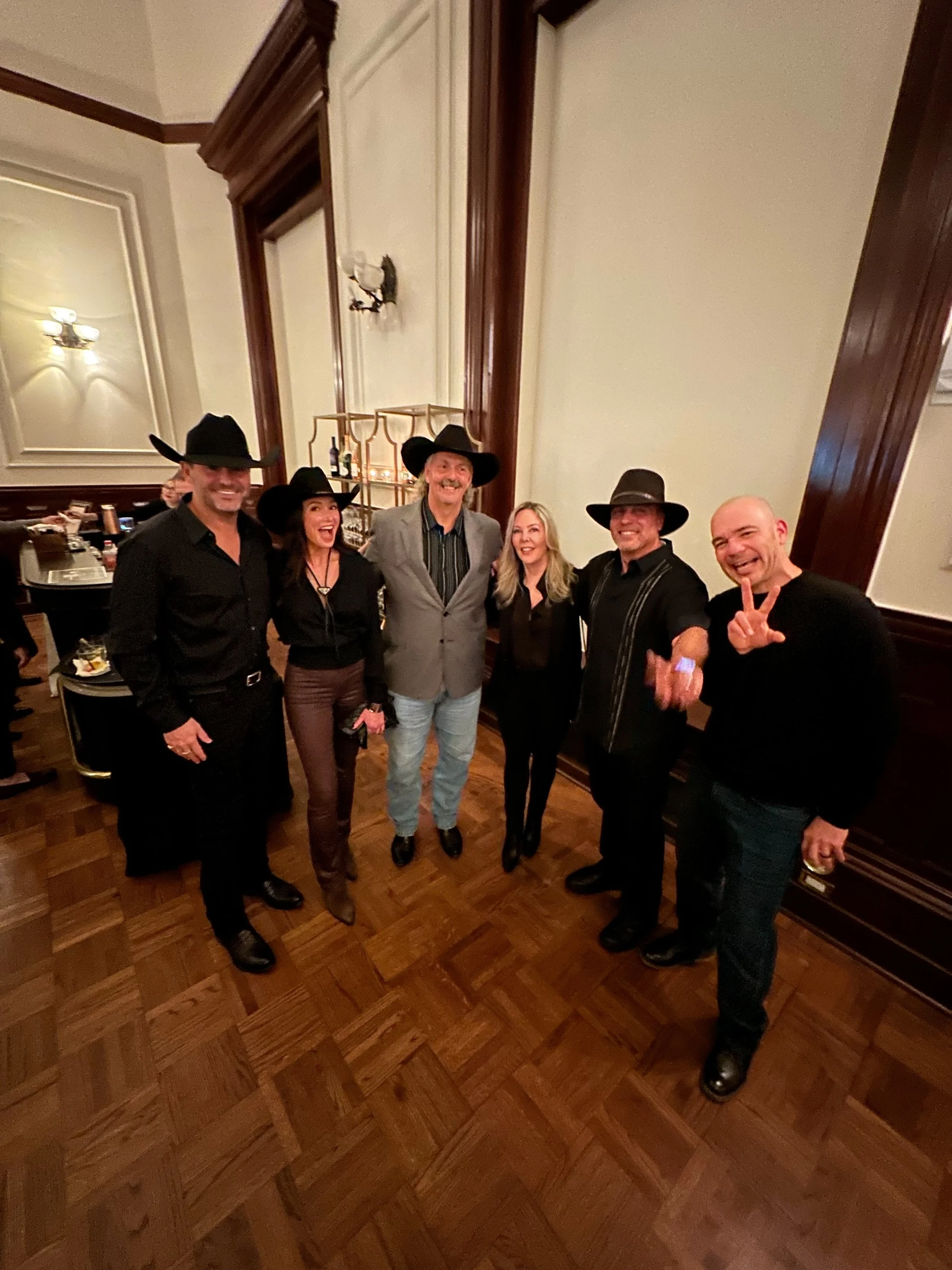 Group of six people in a room, four men and two women, all wearing black hats, with some dressed in black shirts, and smiling or making gestures.