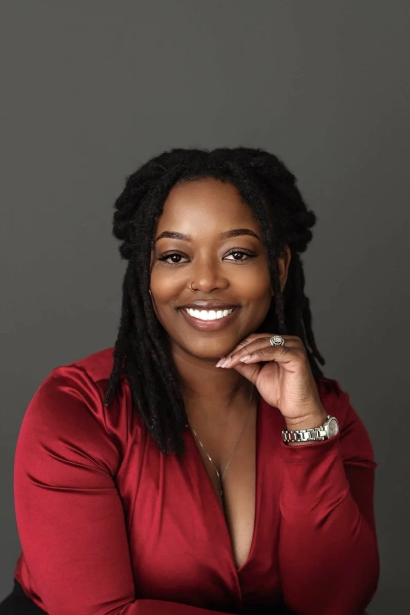 A woman with dark curly hair and a nose ring smiling while wearing a red blouse and jewelry, against a gray background.