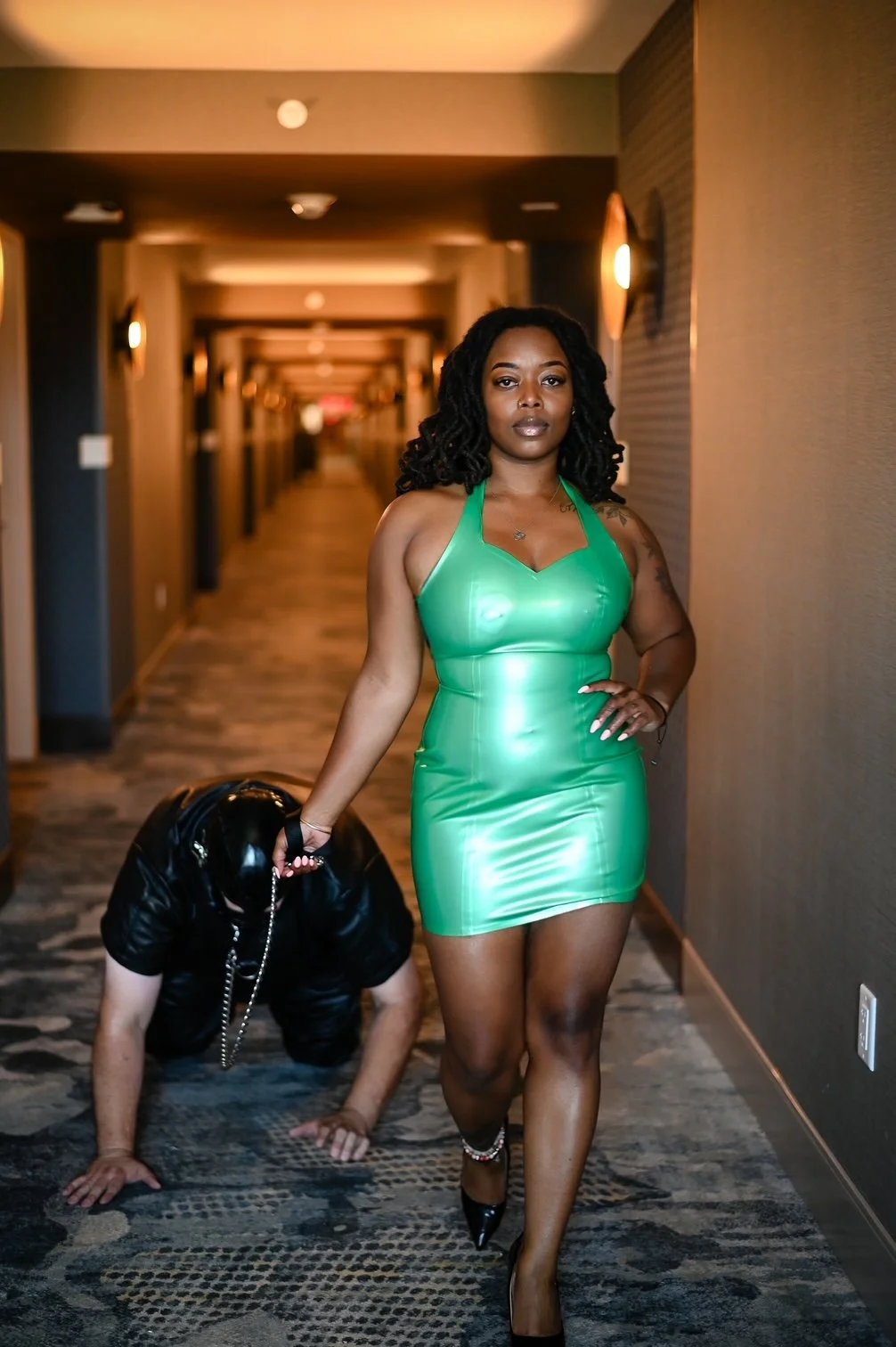 A confident woman in a green latex dress and high heels walks down a hotel hallway with a person dressed in black and on all fours behind her.
