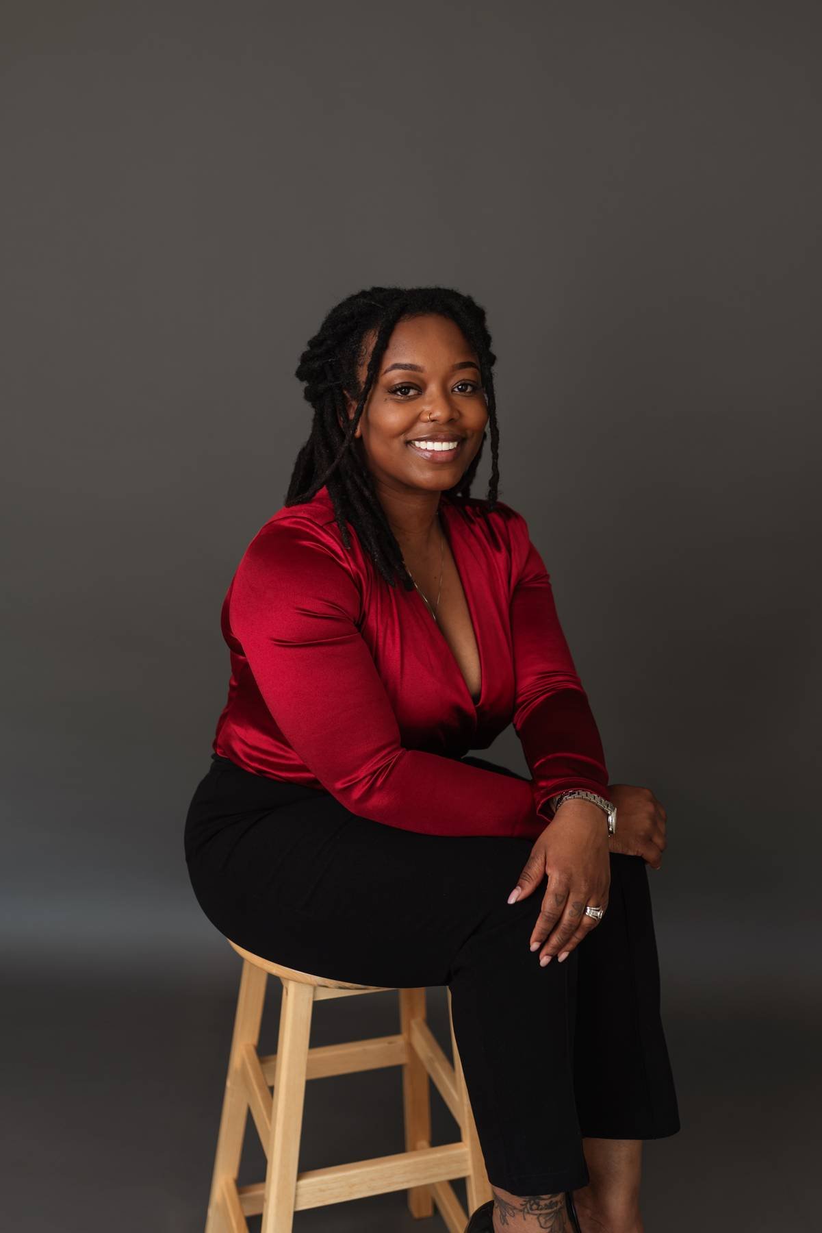 A woman with shoulder-length dreadlocks wearing a red satin blouse and black pants sitting on a wooden stool against a dark gray background, smiling at the camera.