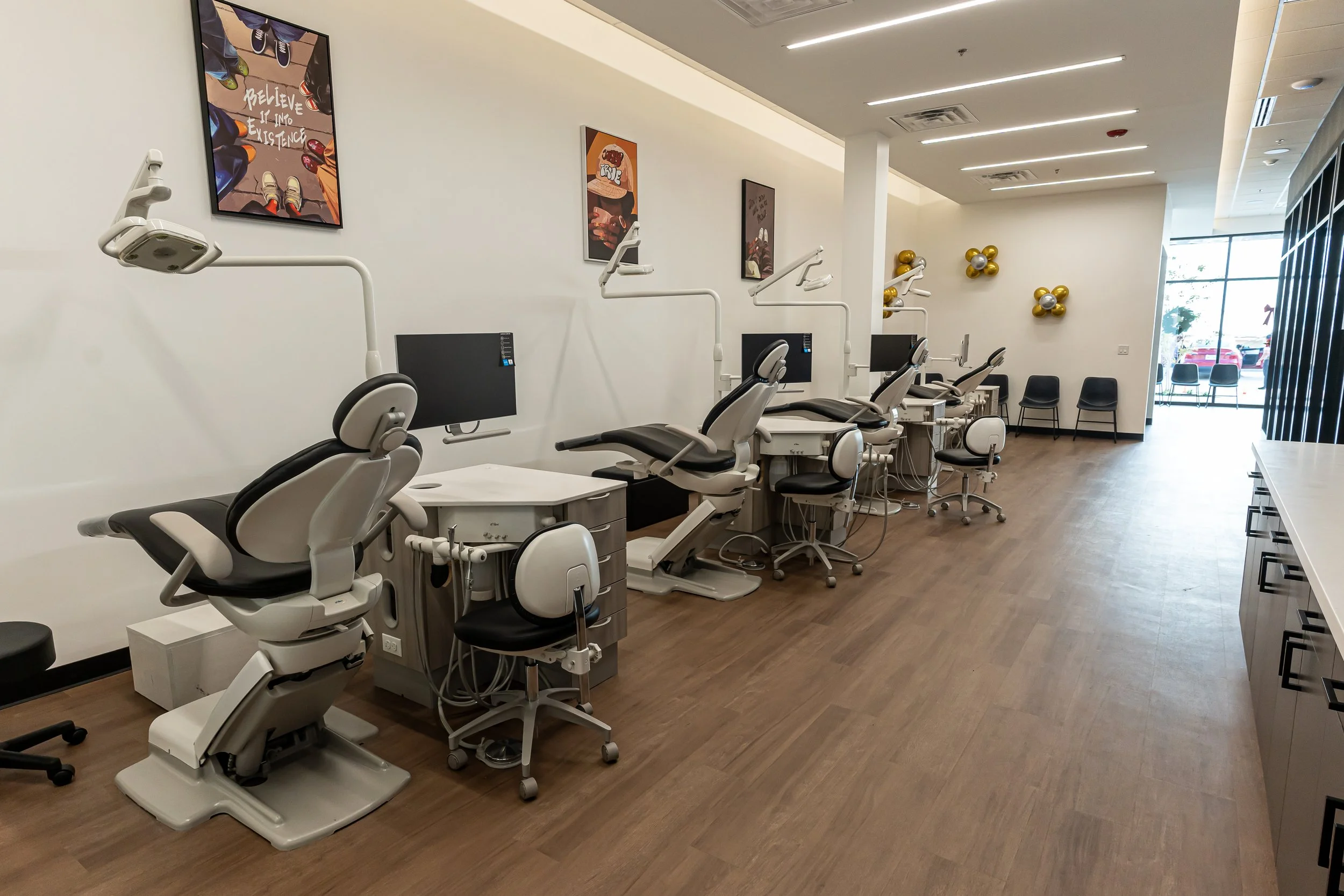 Empty dental clinic room with dental chairs and equipment, black chairs along the wall, artwork and balloons on the wall, large windows at the front.