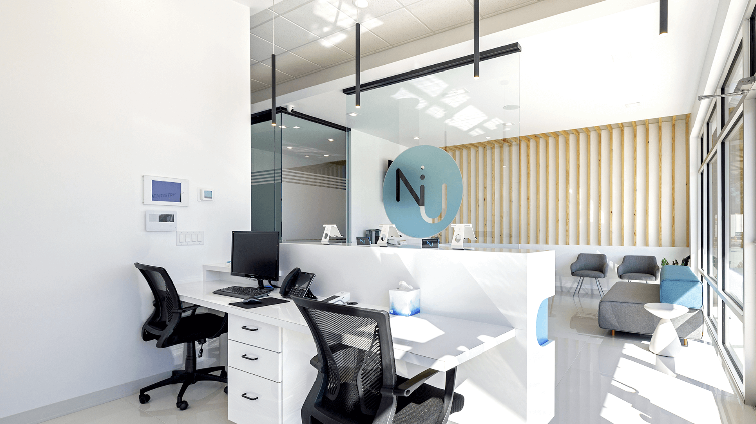 Modern medical office reception area with white walls, glass partition, black chairs, and a waiting area with gray and blue chairs near large windows.
