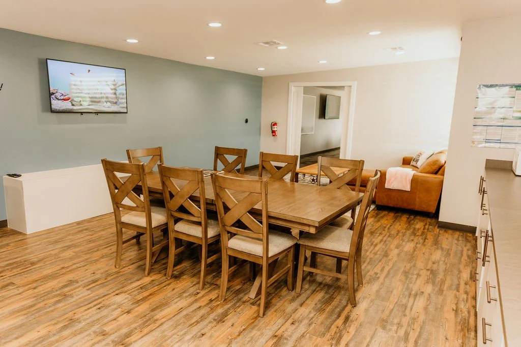 A dining area with a large wooden table and eight matching chairs, hardwood flooring, a wall-mounted TV, and a small sitting area with a tan sofa and blanket in the background.