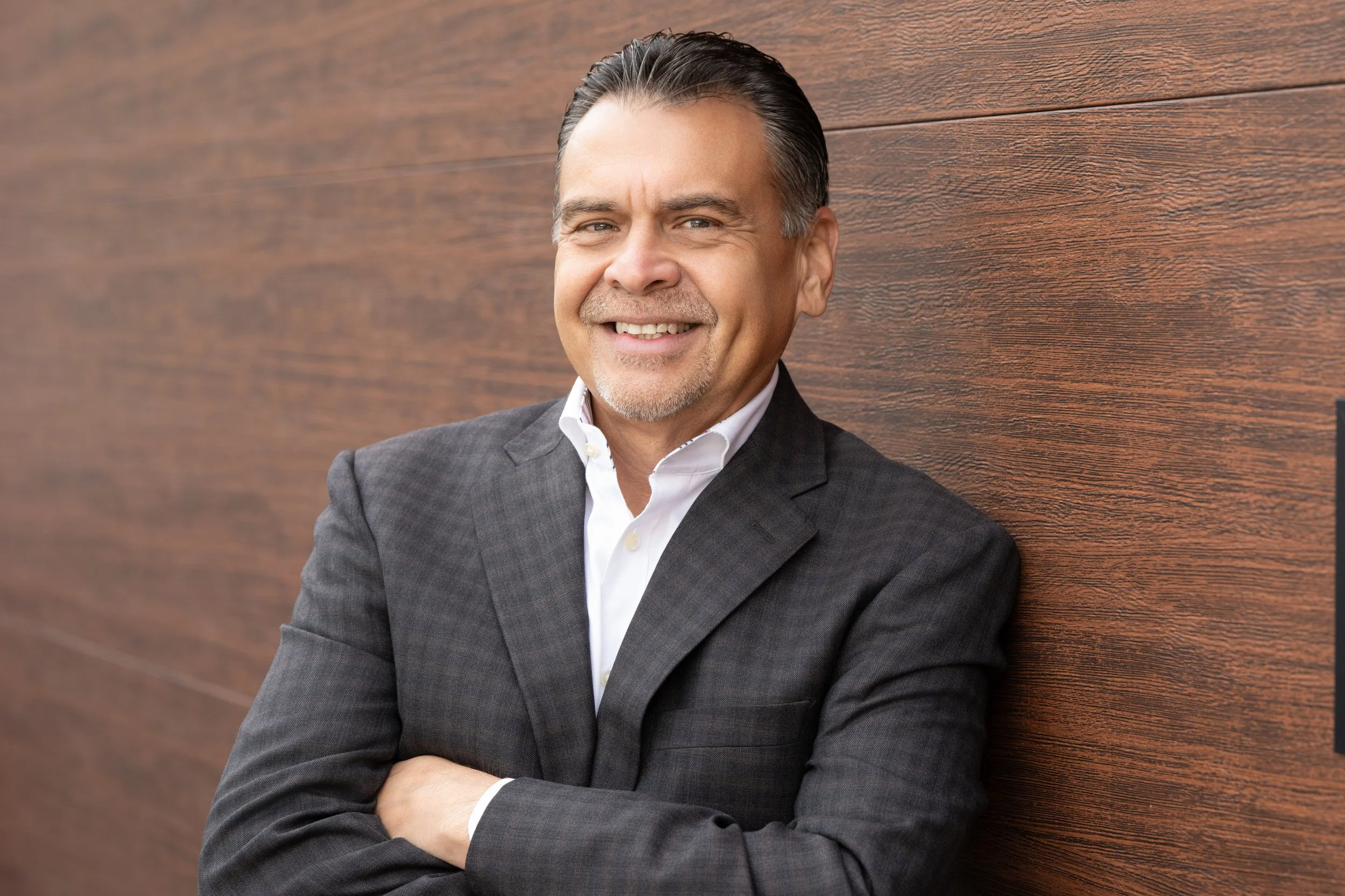 A middle-aged man with dark hair, wearing a dark blazer and white shirt, smiling and leaning against a wooden wall with arms crossed.