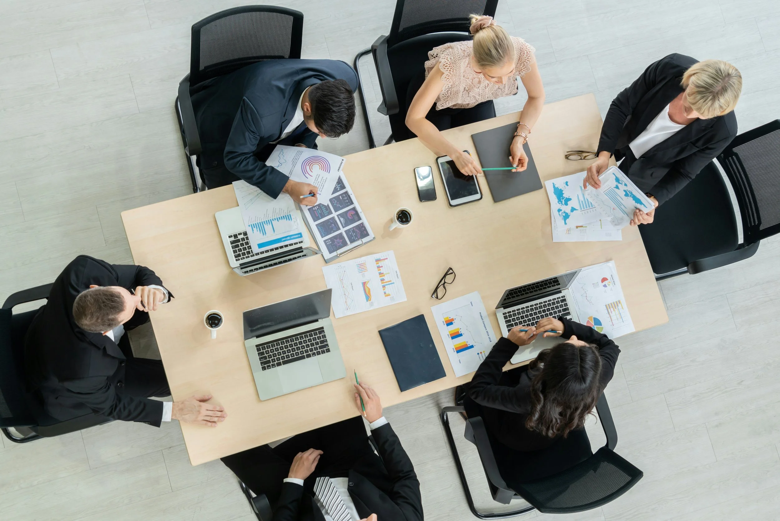 Overhead view of six professionals in a business meeting around a rectangular table, discussing charts, graphs, and documents.