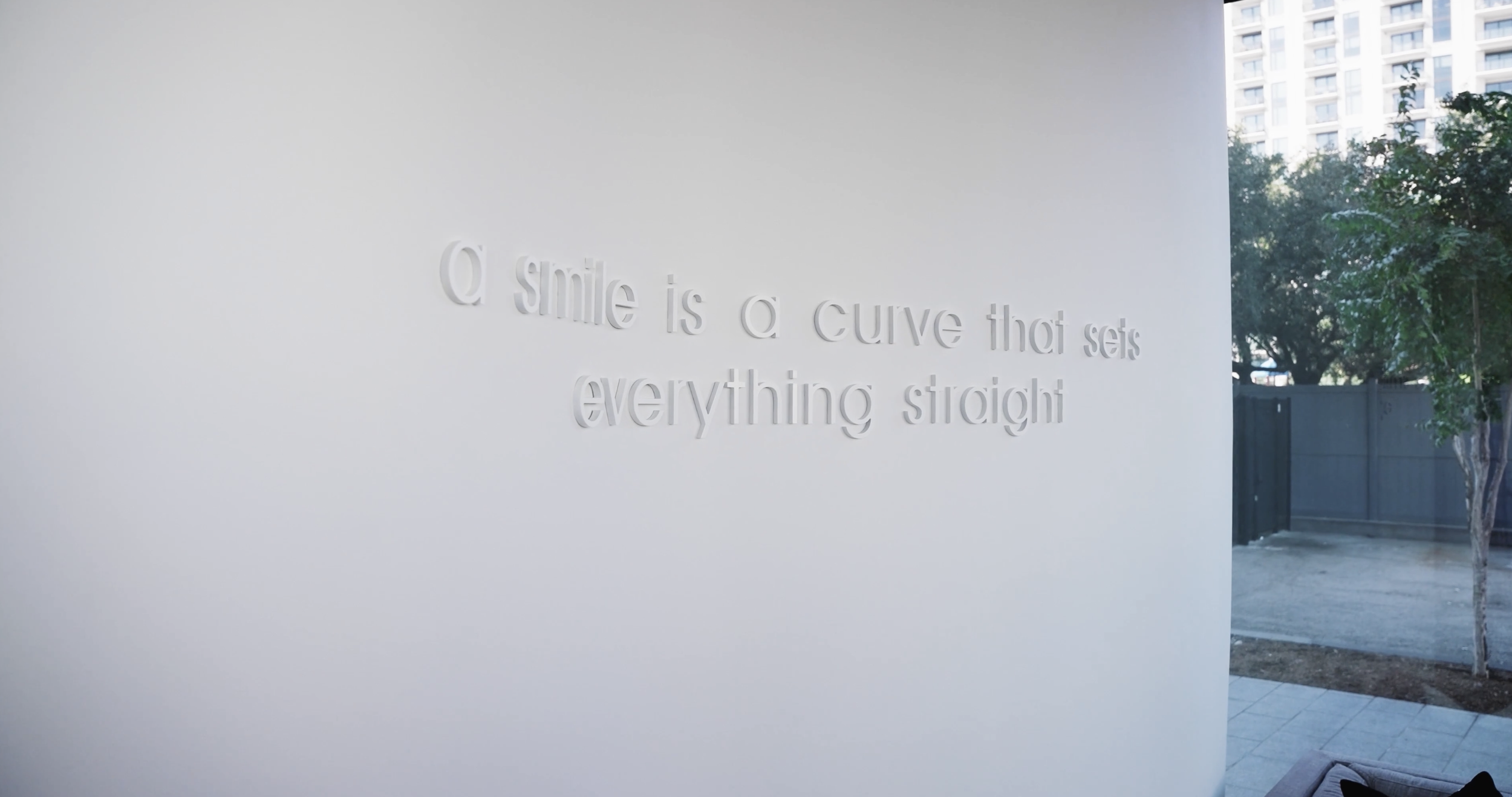 A white wall with a quote that reads: "a smile is a curve that sets everything straight." in raised letters, with trees and a building visible in the background.