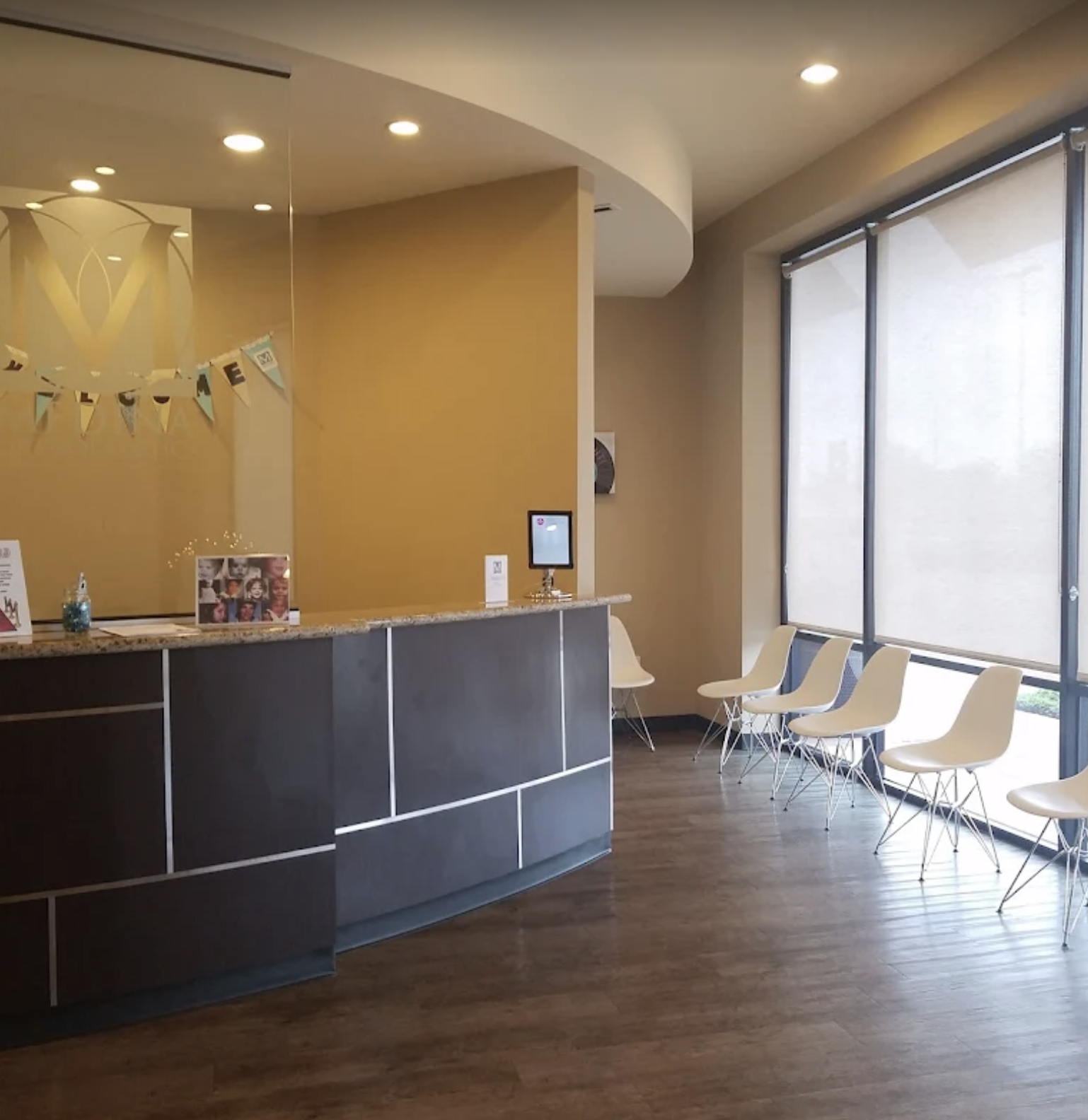 Empty waiting area with a row of white chairs lined up along large windows, a curved reception desk with a granite top, and a yellow wall with festive decorations and photos displayed on the counter.