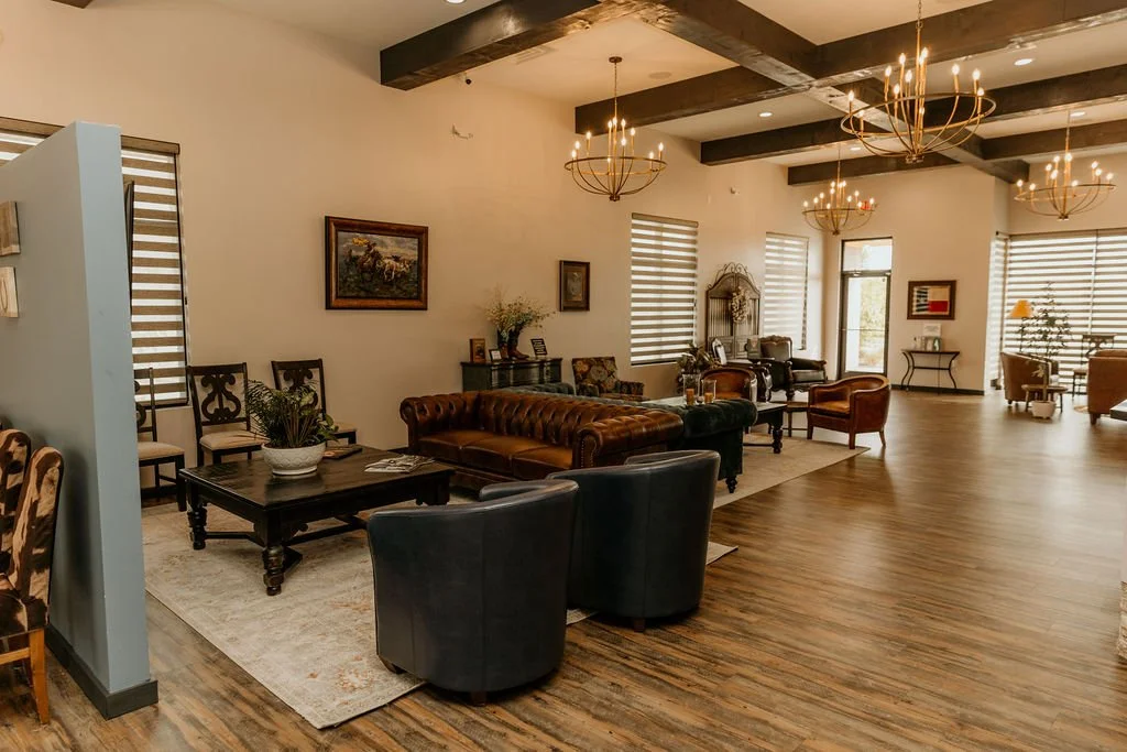 Modern office space with large windows, glass desk, black leather chairs, a wall-mounted TV, and greenery outside.