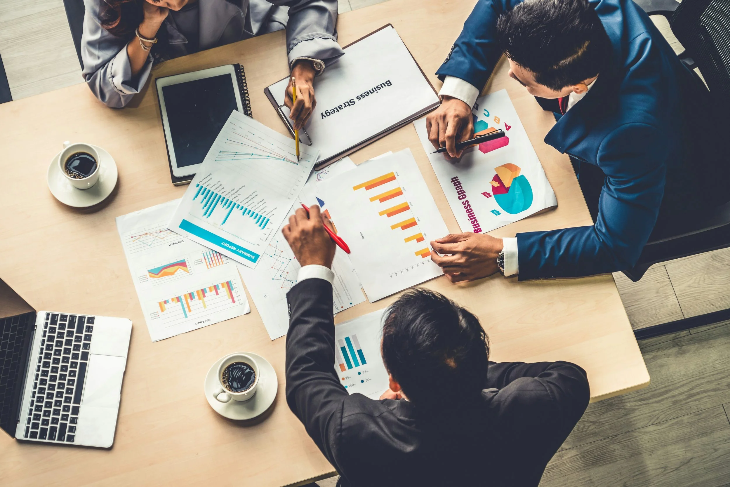 Three business professionals sitting around a wooden table discussing, with various financial charts, graphs, a laptop, notebooks, and cups of coffee on the table.
