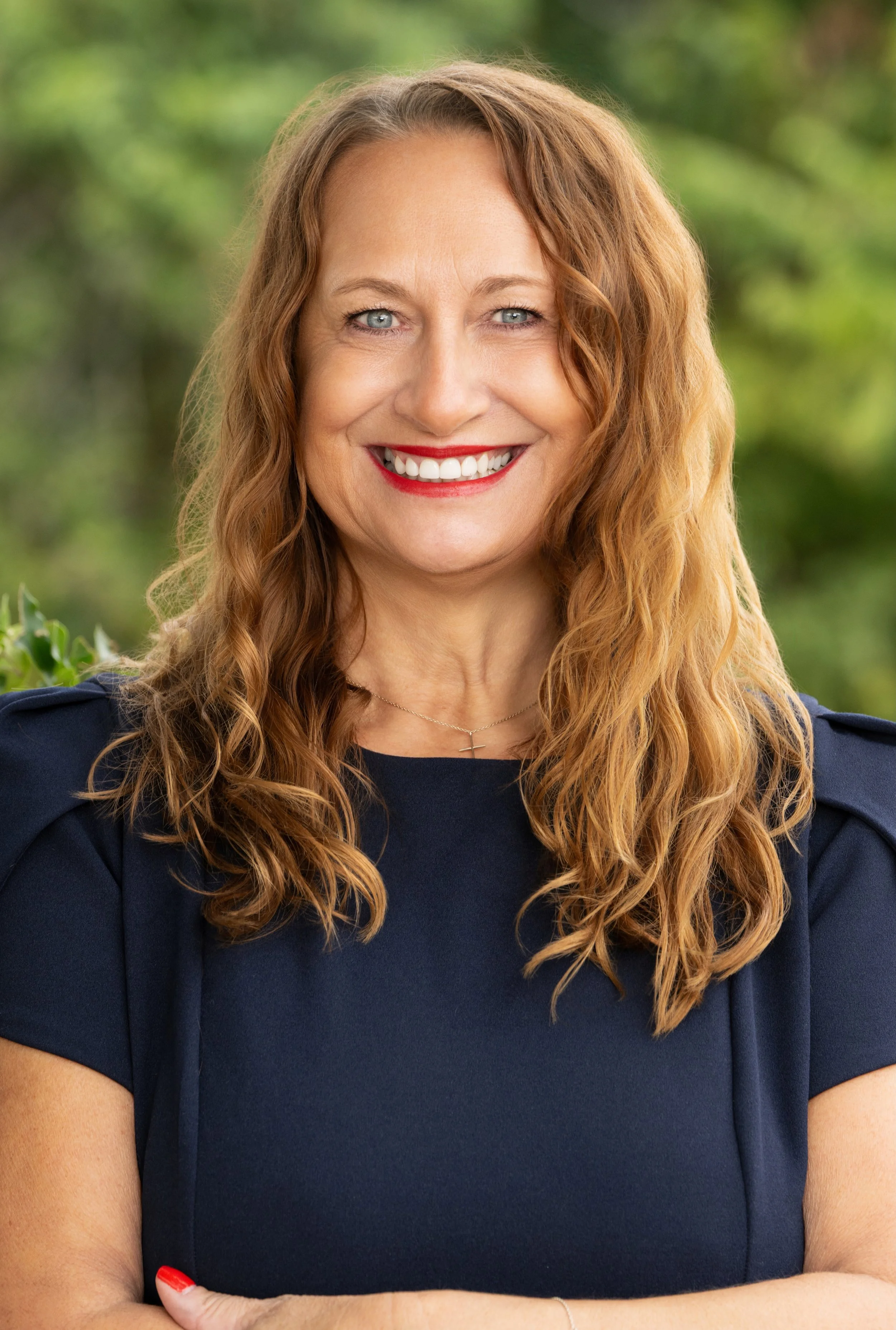 Smiling woman with long red wavy hair wearing a navy blue top, outdoors with green trees in the background.