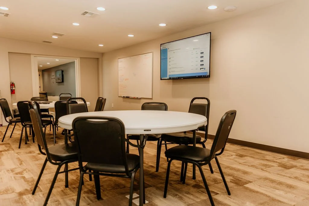 A conference room with round tables and black chairs, a whiteboard, and a mounted TV screen showing a messaging app, with beige walls and ceiling lights.