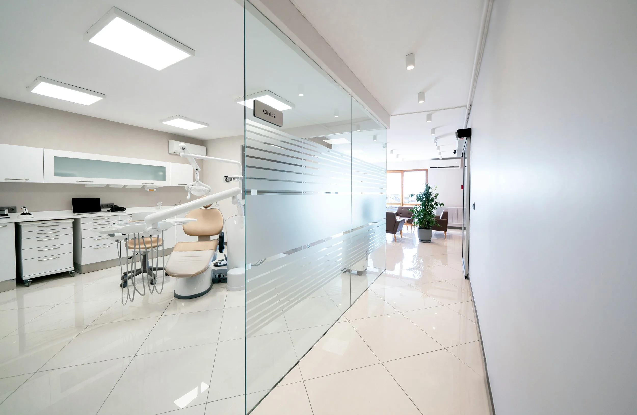 Inside a modern dental clinic with a dental chair and equipment behind a glass wall, and a waiting area with chairs and a potted plant near a large window.