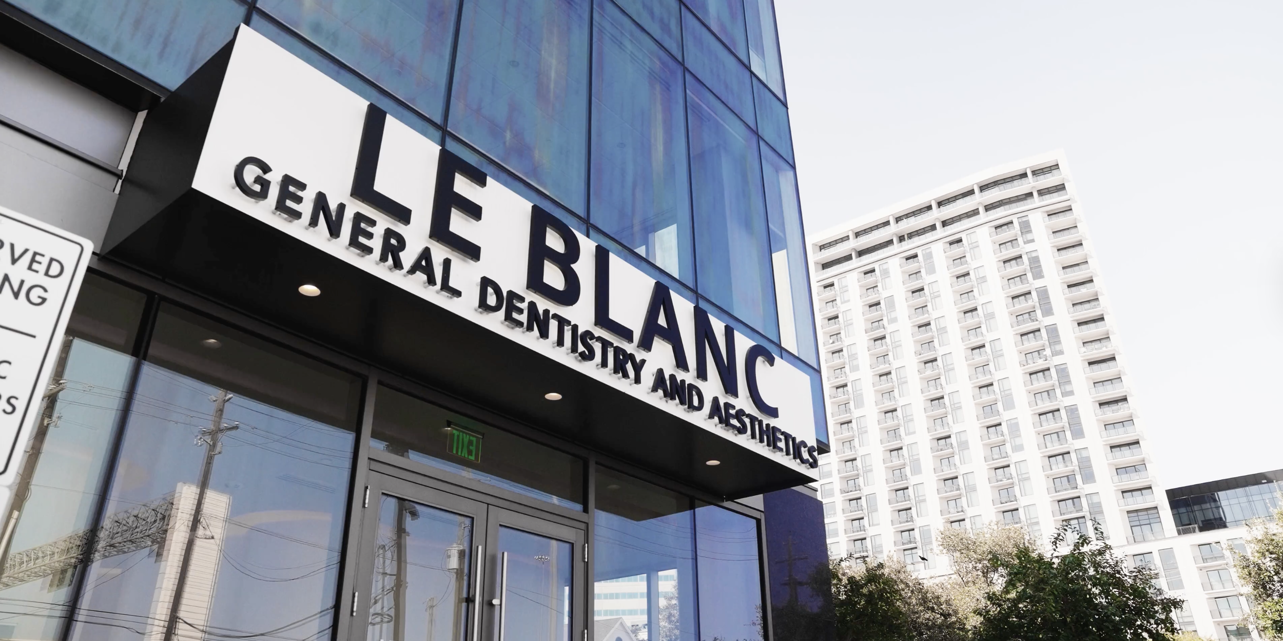 Exterior view of a modern building with a sign that reads 'LEBLANC, General Dentistry and Aesthetics' in bold black letters. The building has blue-tinted glass windows, and there is a high-rise apartment building in the background. A green exit sign is visible above the door.