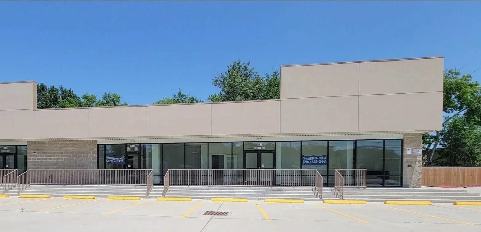 A vacant commercial storefront with large glass windows and doors, a ramp and stairs at the entrance, yellow parking lot lines, and a blue sky overhead.