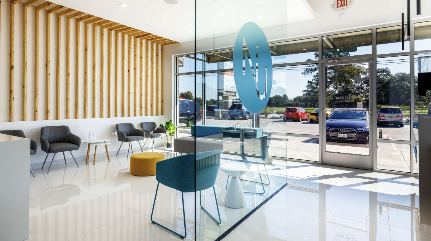 Modern lobby area with gray chairs, a yellow ottoman, a green plant, glass walls, and a parking lot visible outside.
