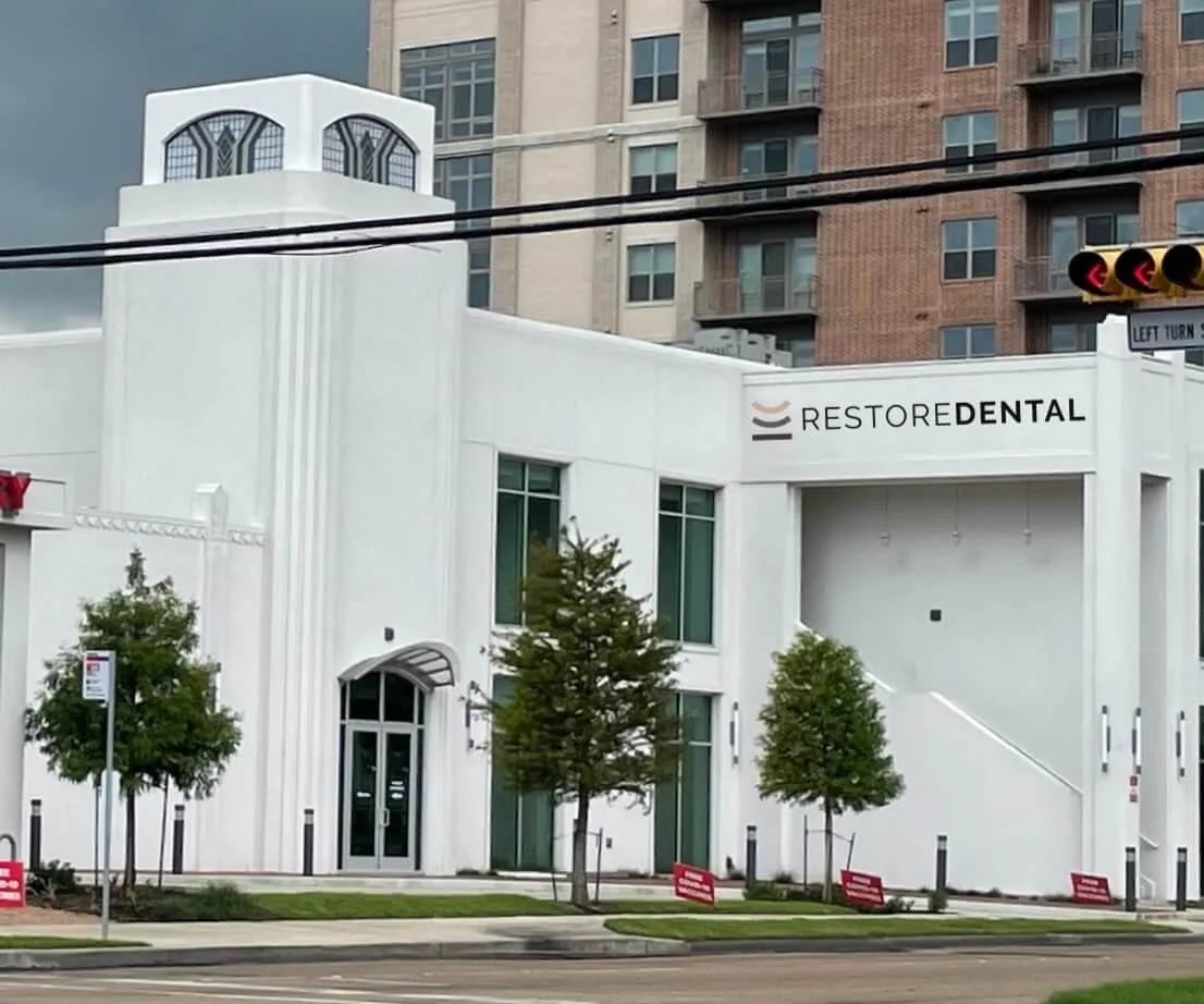 White building with a sign that reads Restore Dental, surrounded by small trees and parked signs, in an urban area with high-rise apartment buildings in the background.