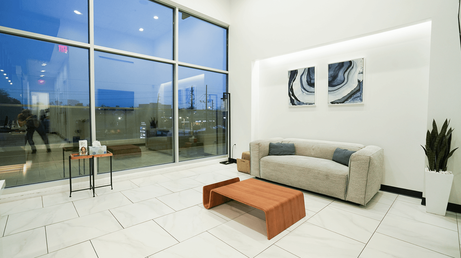 Modern lobby with large windows, white walls, a beige sofa with blue pillows, a wooden coffee table, abstract wall art, and a tall potted plant.