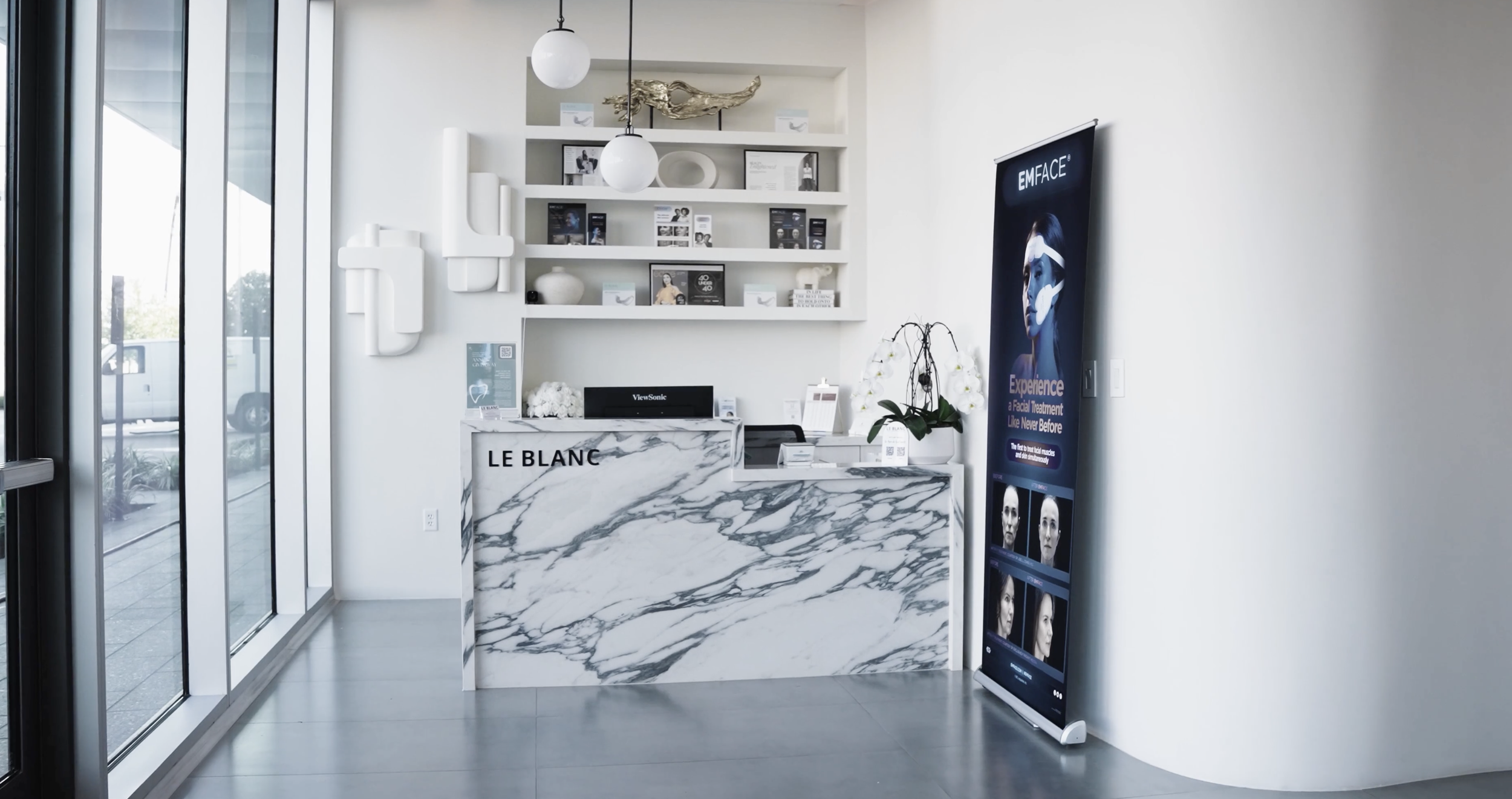 Modern reception area with a white marble desk, white shelves with decorative objects, a vibrant blue poster stand, and large glass windows