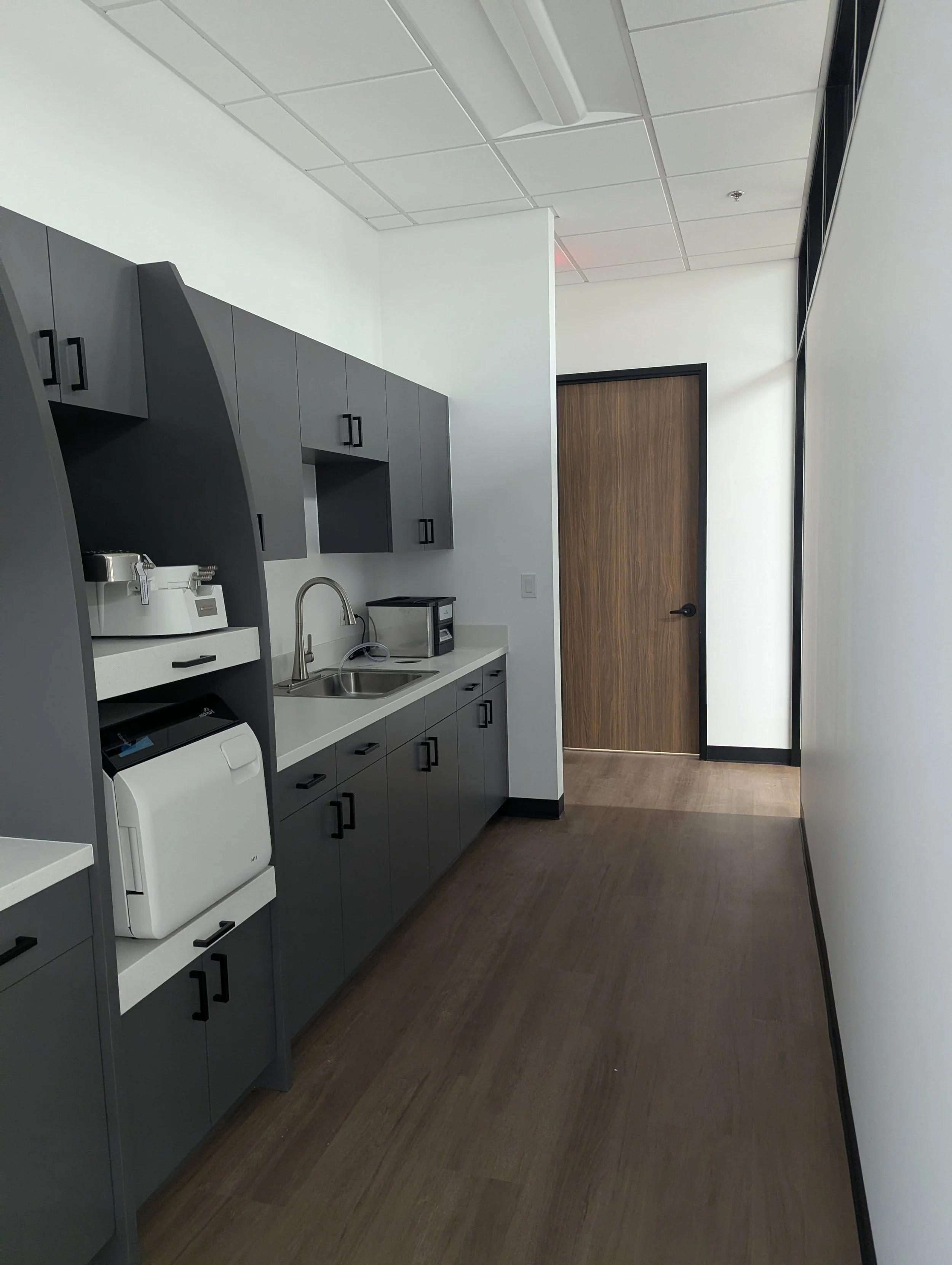 An office kitchenette with gray cabinets, a white countertop, a sink with a faucet, and various small appliances including a microwave, toaster, and a white machine, with a closed wooden door in the background and a white wall on the right side.