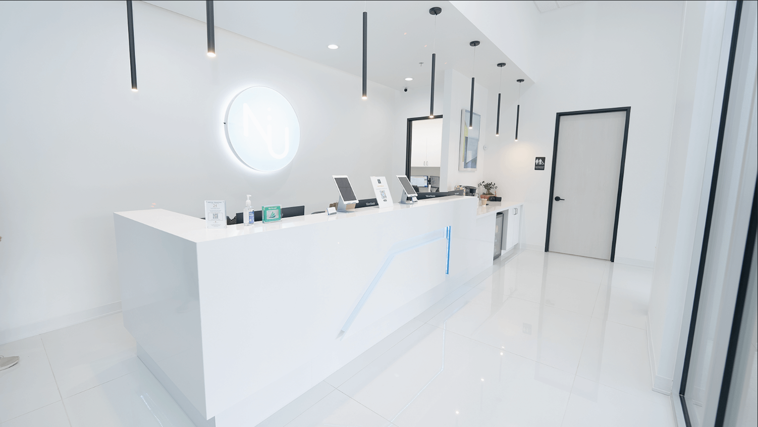 Modern and minimalist reception area with a white counter, digital displays, disinfectant hand sanitizer, and a neon sign with the letters 'NU' on the wall. The space has white walls, black accents, and hanging black pendant lights.